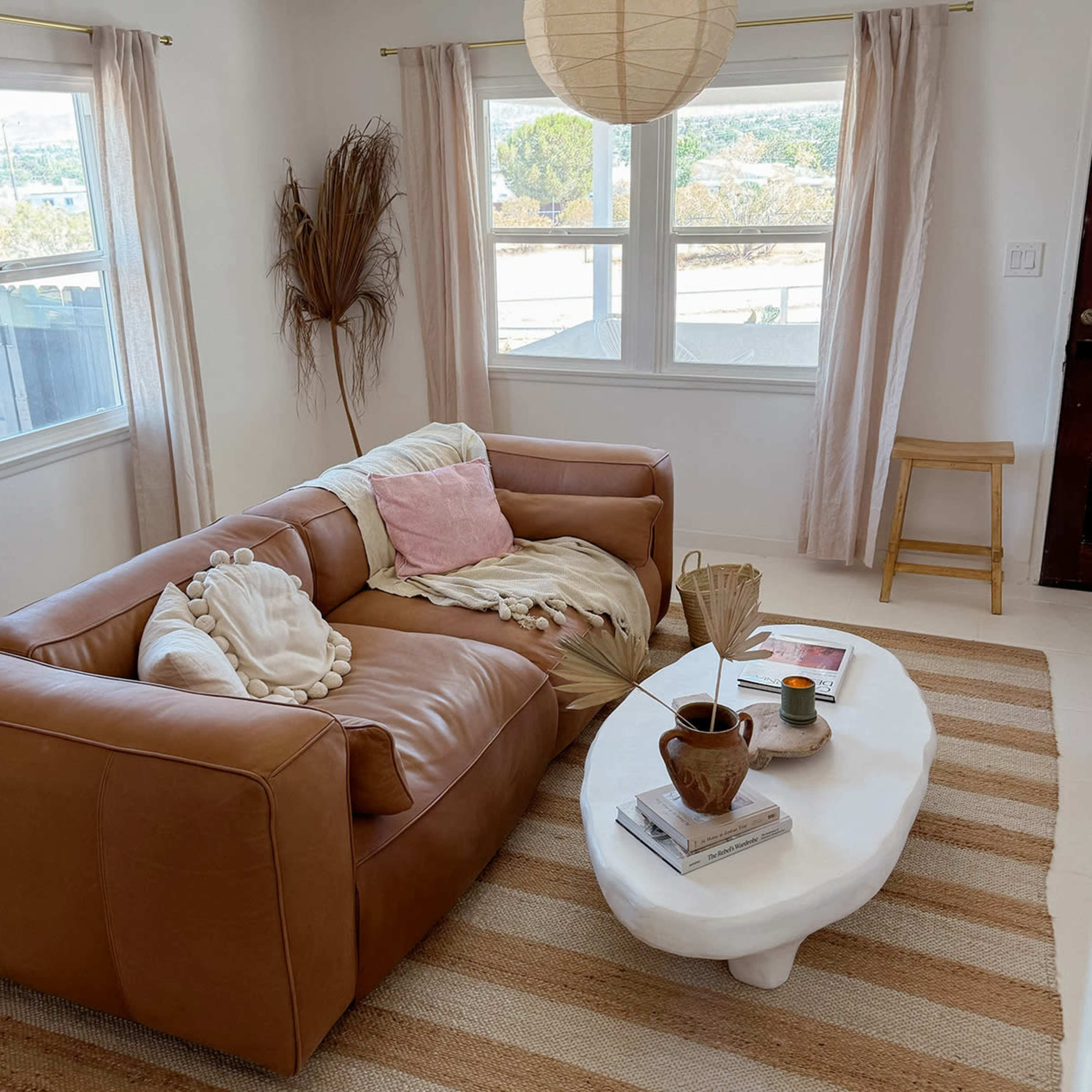 A cozy living room featuring a brown leather sofa, a white coffee table, and natural light streaming through sheer curtains.