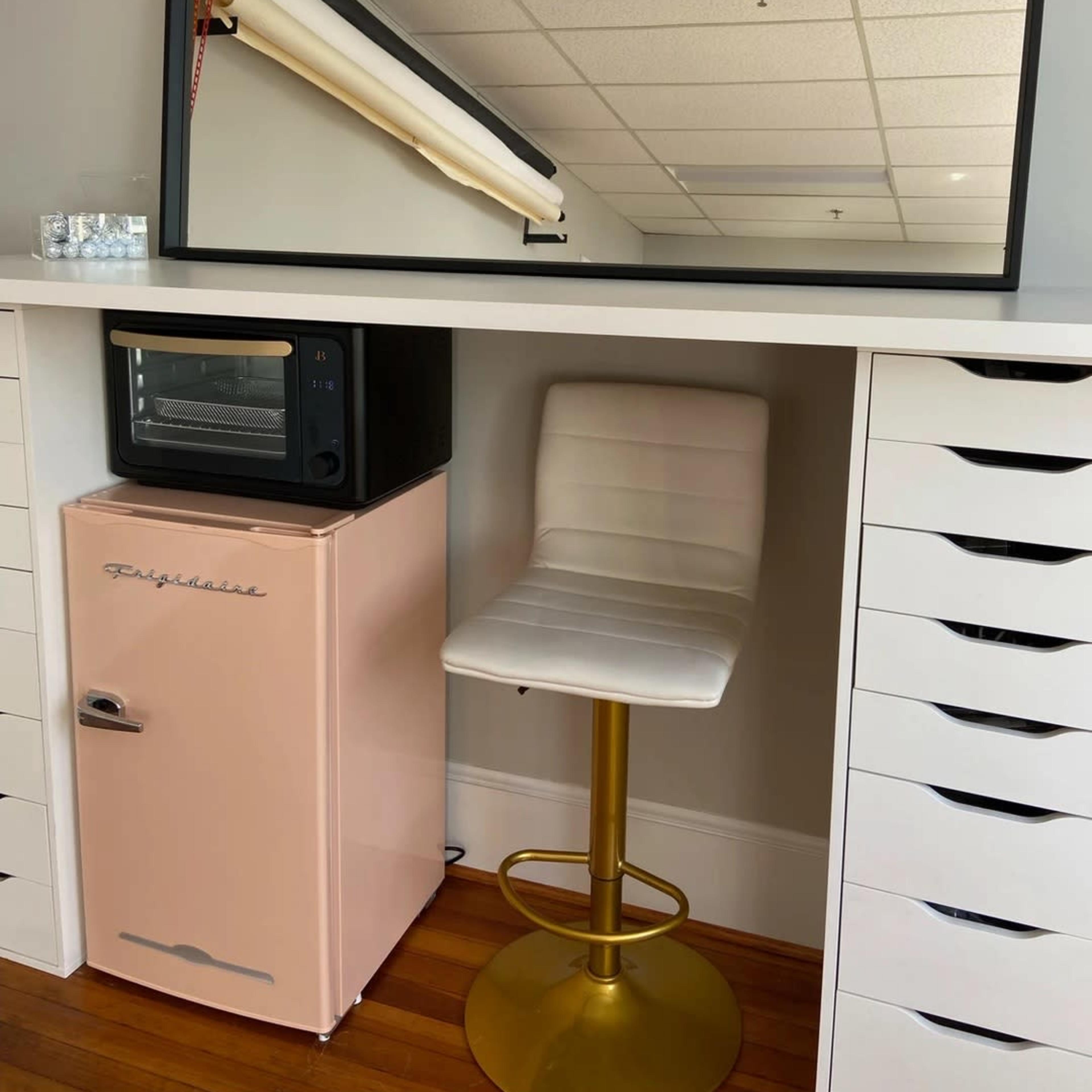 A mirror hangs above a countertop with a pink refrigerator, a black microwave, and a gold bar stool.