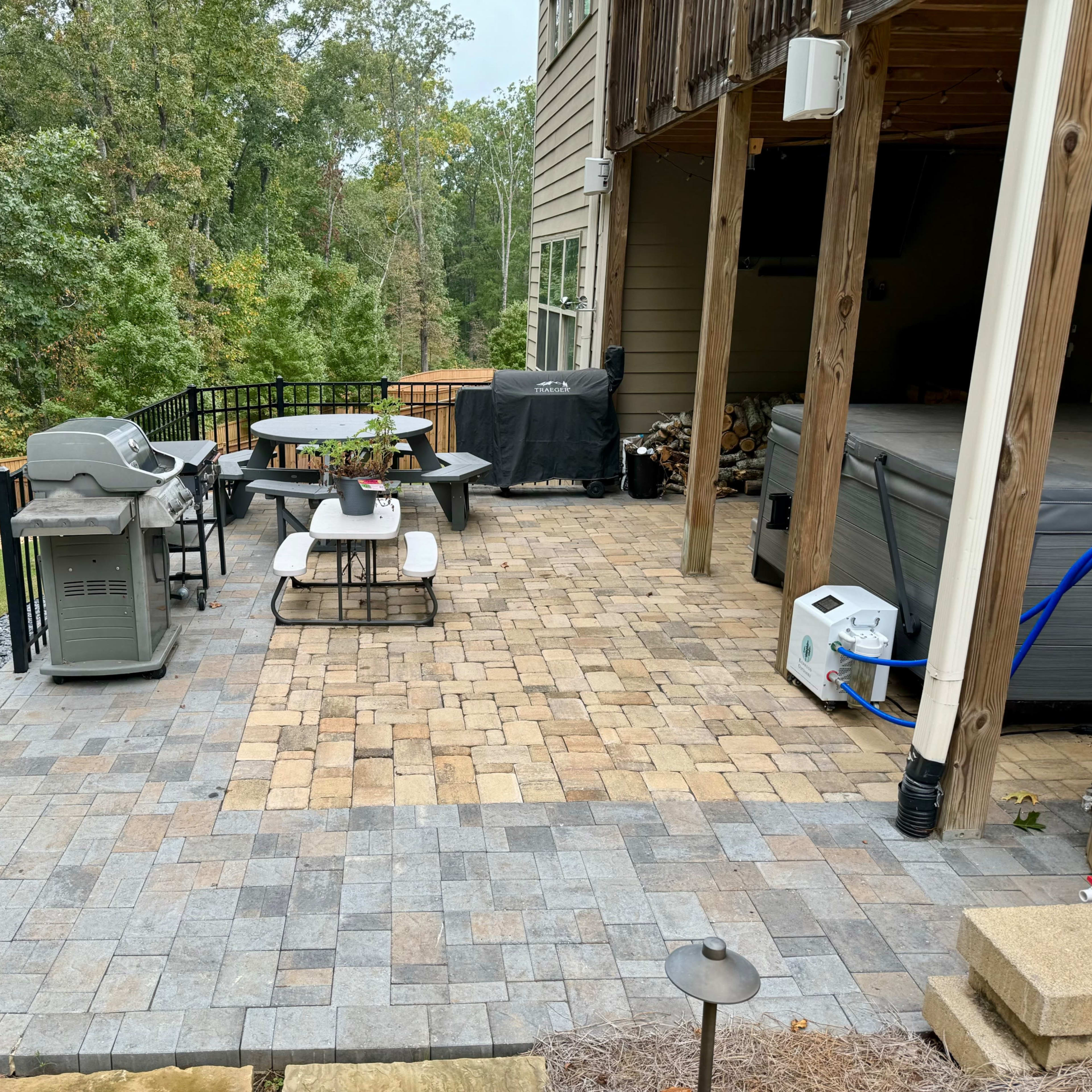 A stone patio area with a table and chairs, a grill, and a hot tub, surrounded by trees and a grassy lawn.