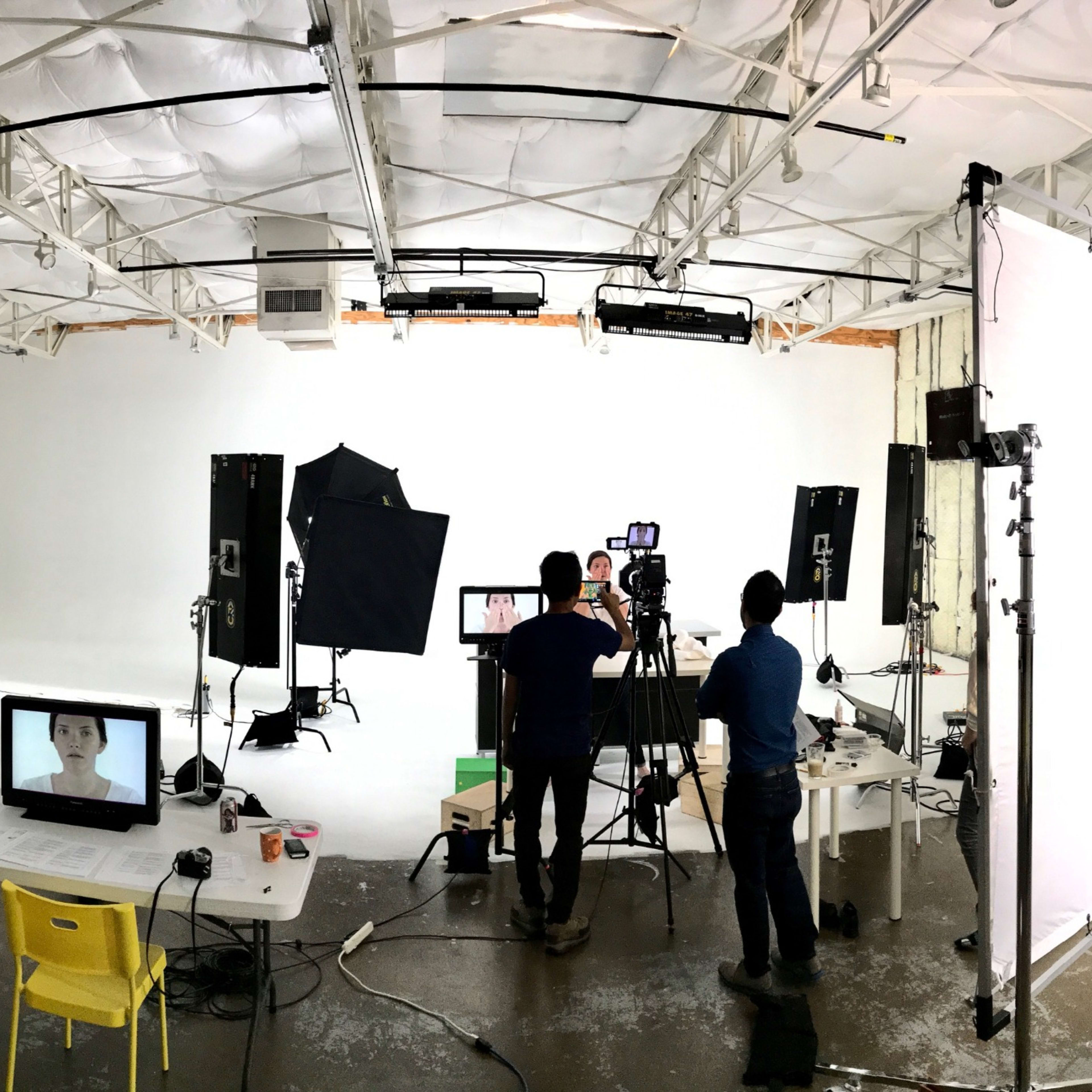 A photo shows a spacious filming studio with lighting equipment, cameras, and a production team working on a set surrounded by various chairs and tables.