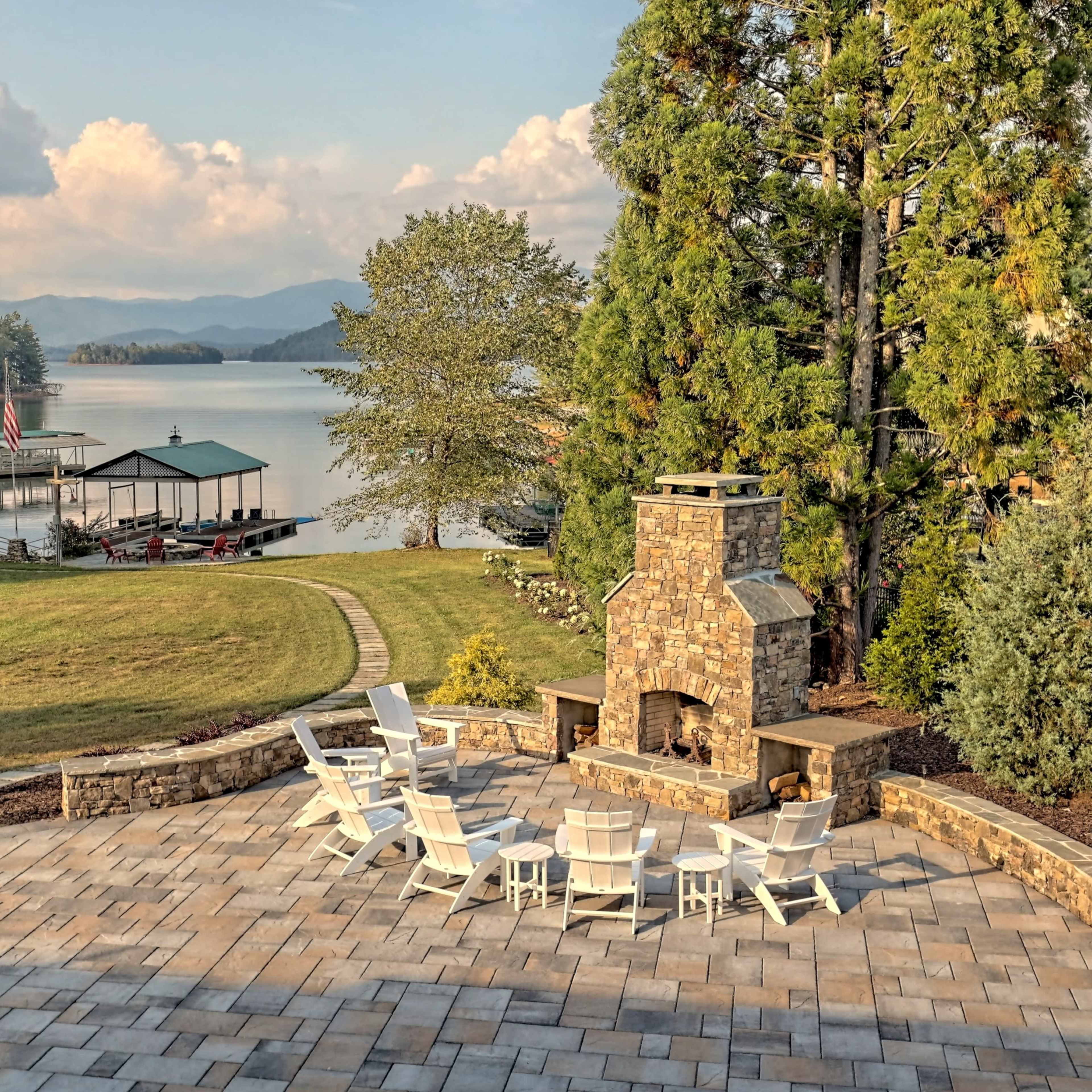 A lakeside patio with white chairs arranged around a stone fireplace, overlooking a calm lake and distant mountains.