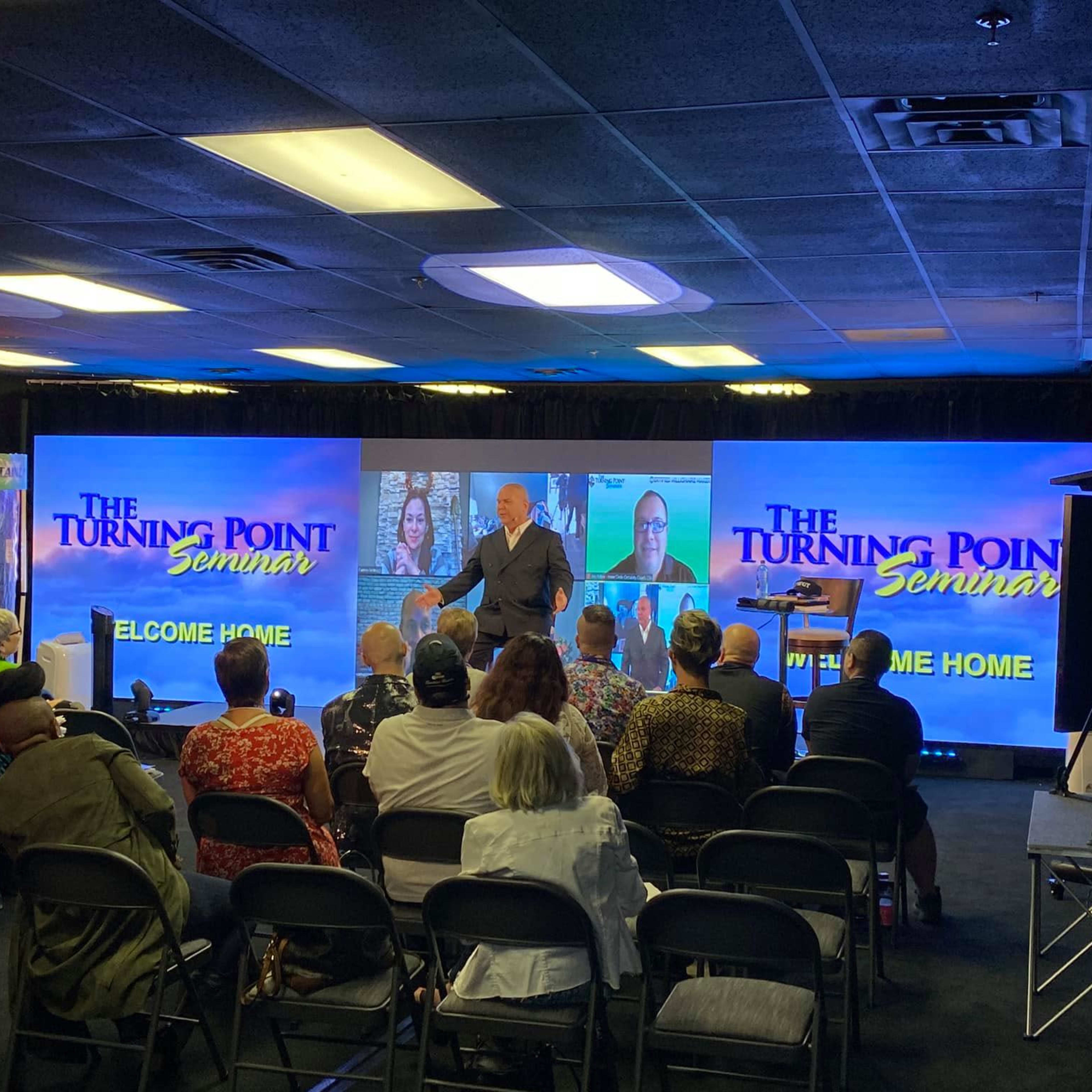 A speaker addresses an audience seated in front of large screens displaying "The Turning Point Seminar" in a conference setting.