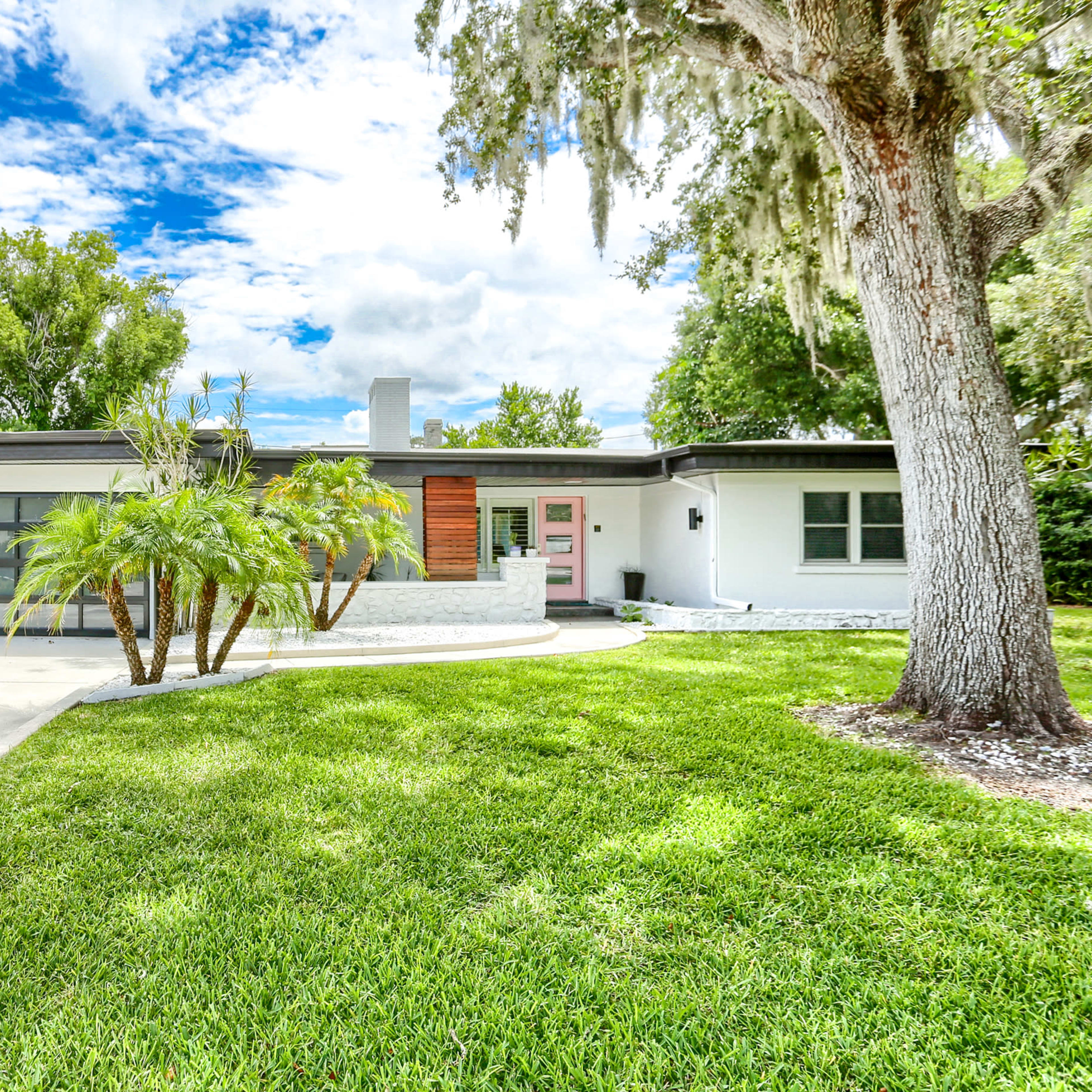 A modern single-story house features a landscaped front yard with palm trees and a driveway leading to a glass garage door.