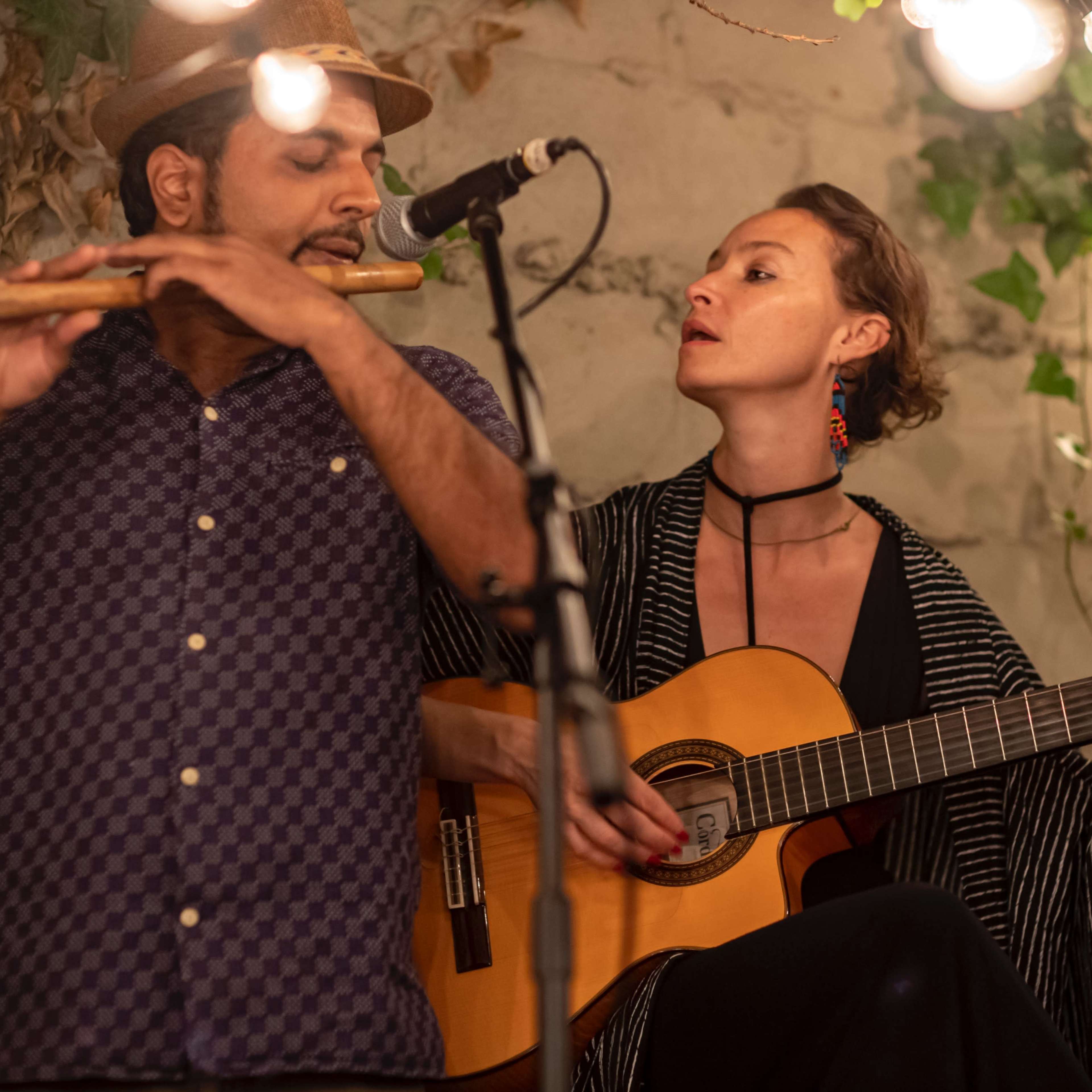 A man plays a flute while a woman strums a guitar, both performing under hanging lights in a greenery-adorned setting.