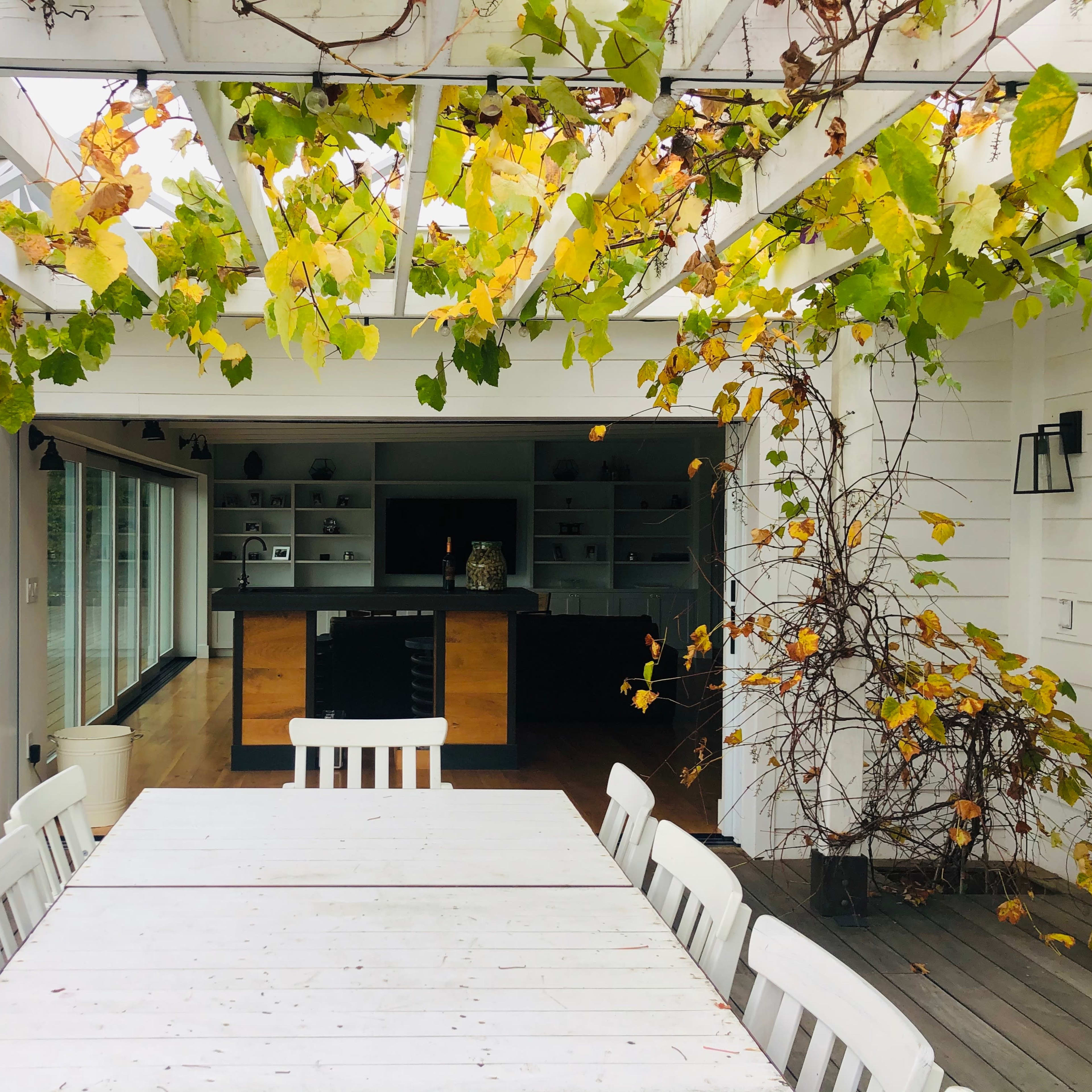 A white outdoor table is set under a pergola adorned with grapevines, overlooking a modern kitchen and a swimming pool.
