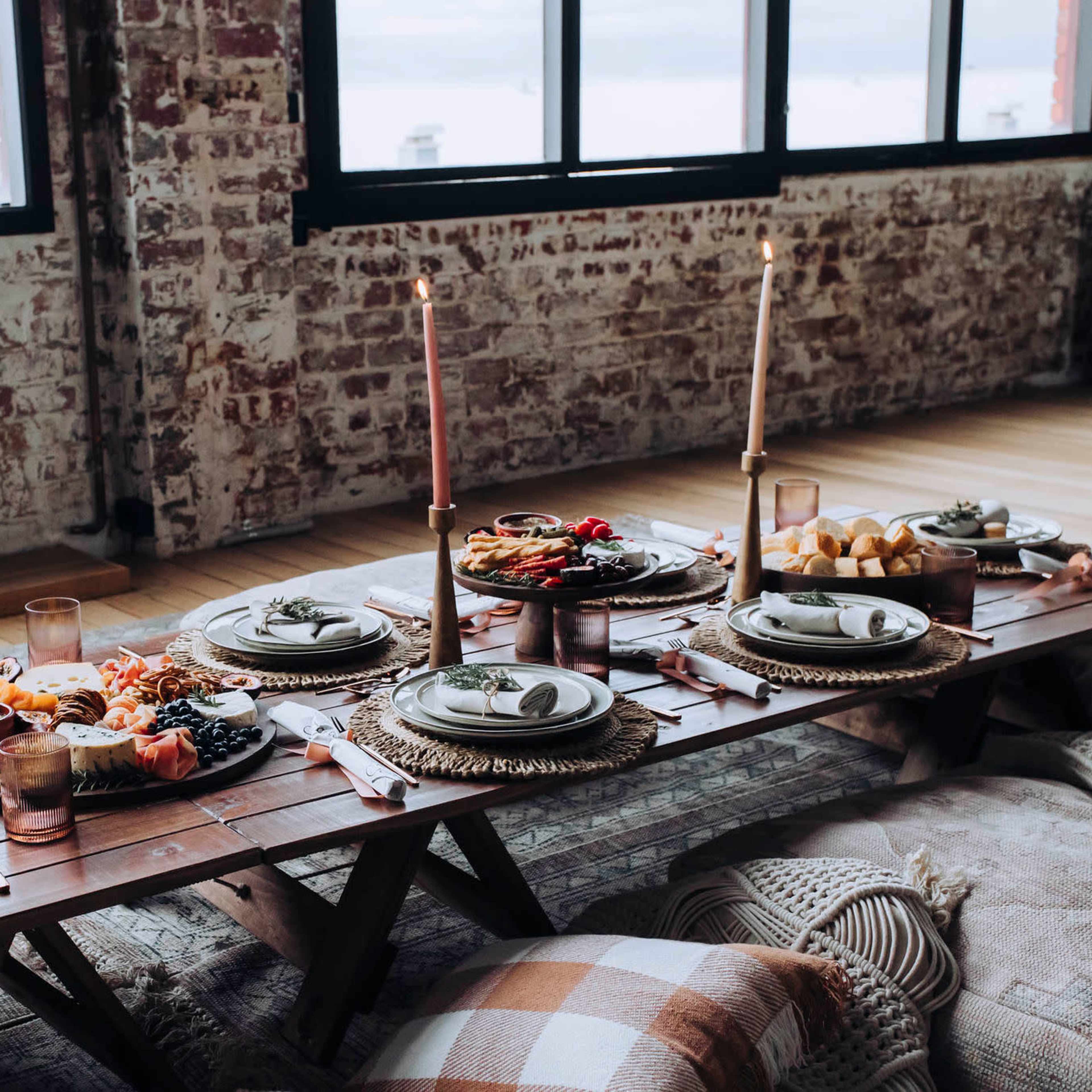 A long wooden table is set with plates, candles, and an array of food in a well-lit room with large windows.