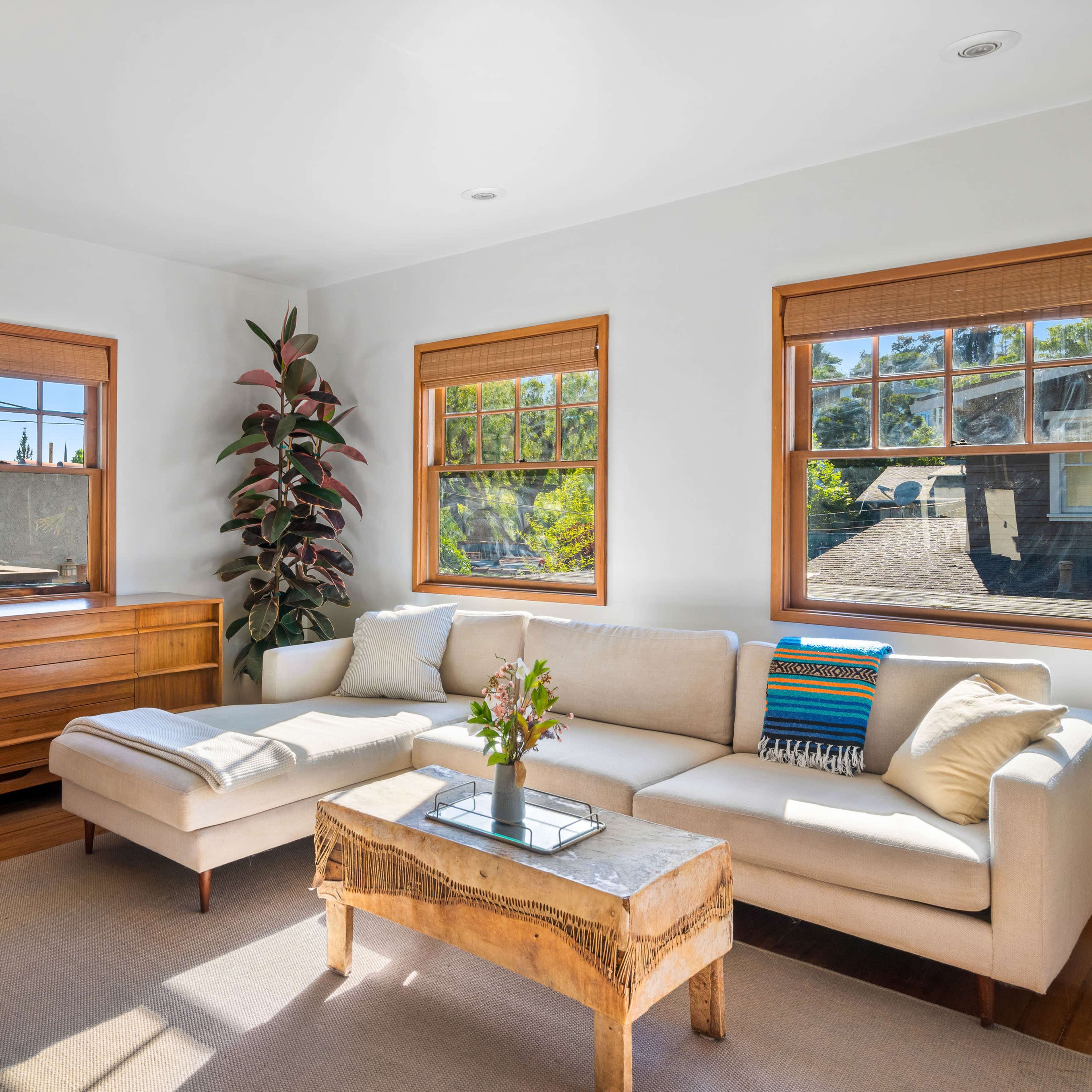 A light-filled living room features a beige sectional sofa, a wooden coffee table, and large windows decorated with bamboo shades.