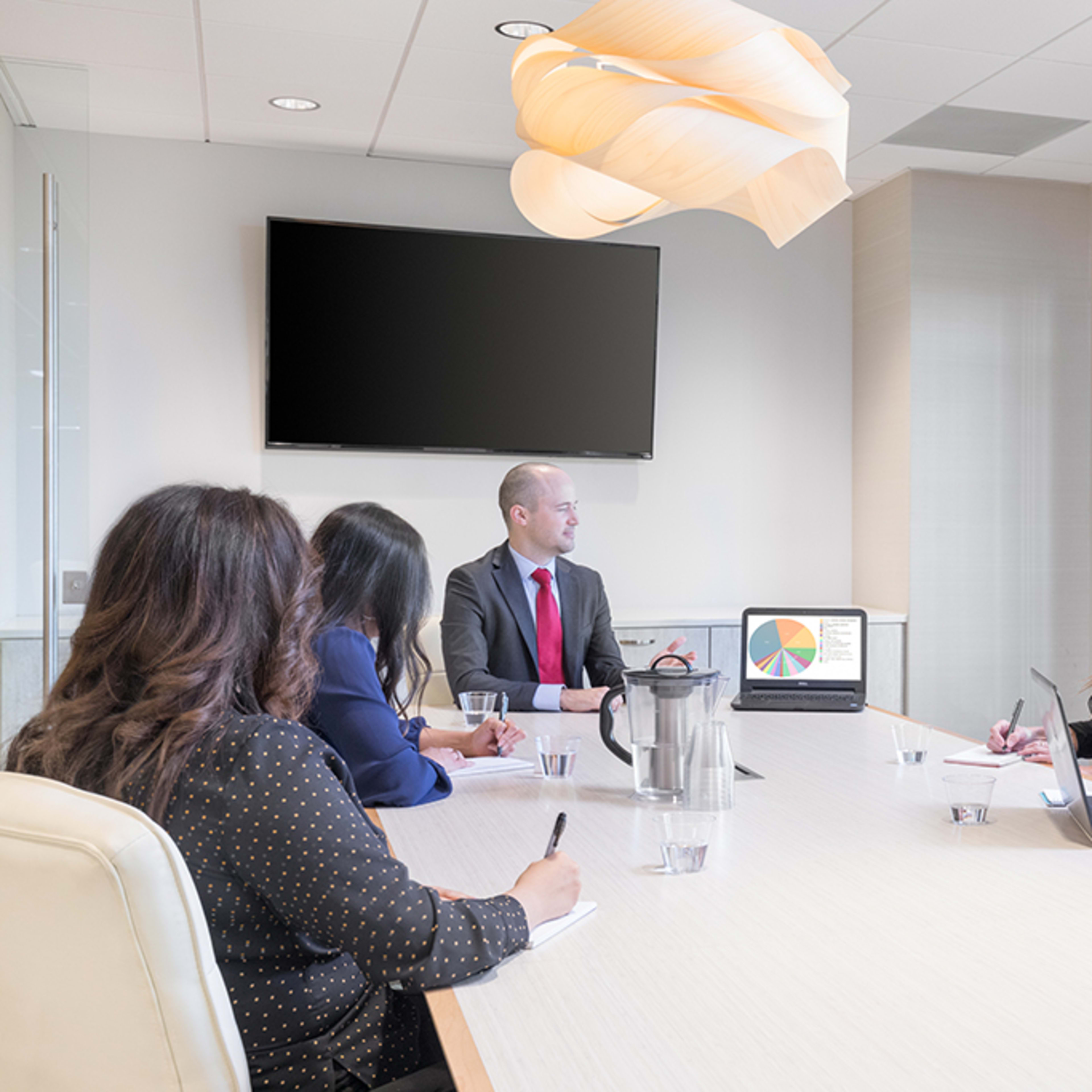 A group of five professionals sits around a conference table, with one man presenting to the others using a pie chart displayed on a laptop.