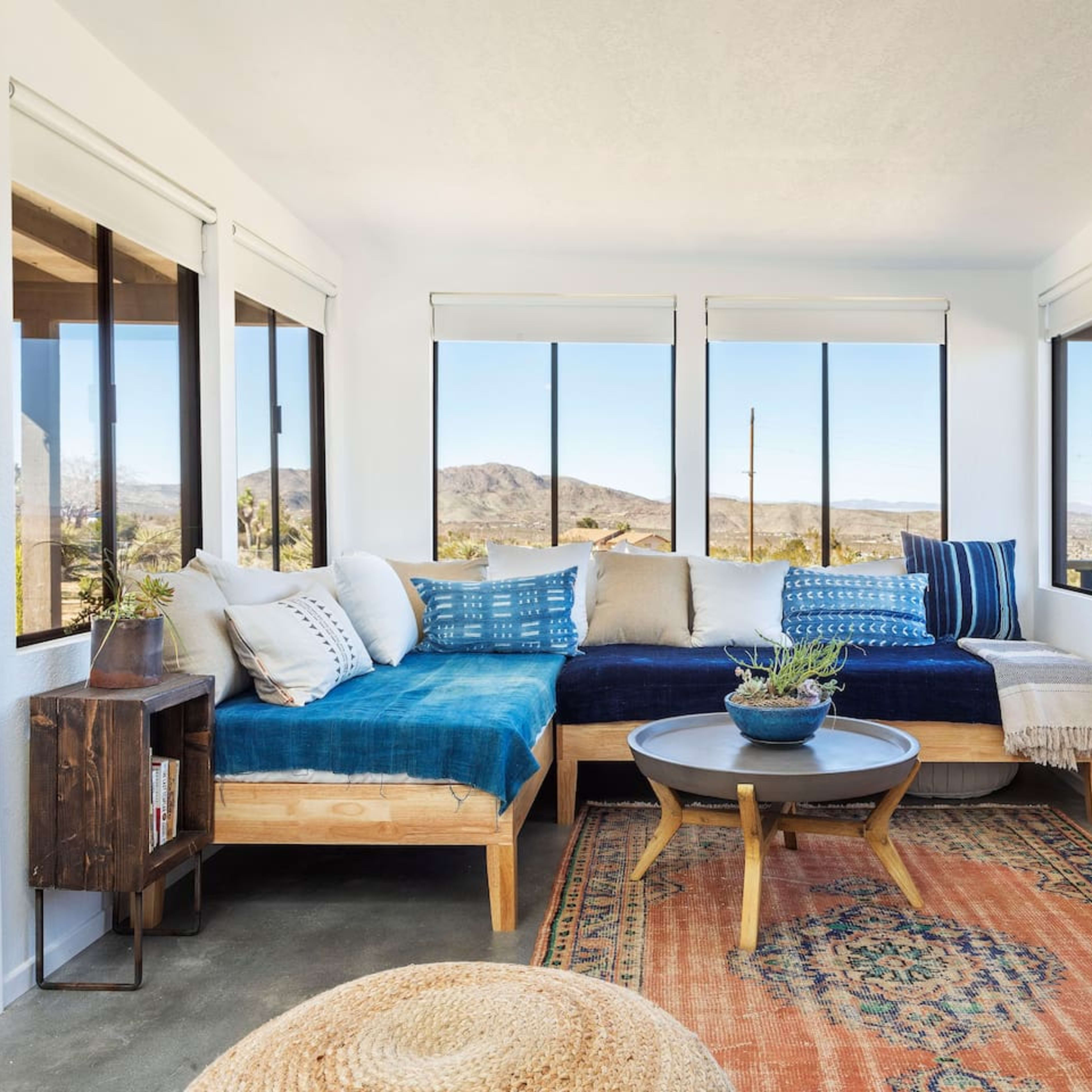 A bright sunroom features a blue and white sectional sofa, large windows with mountain views, and a circular wooden coffee table on a patterned rug.