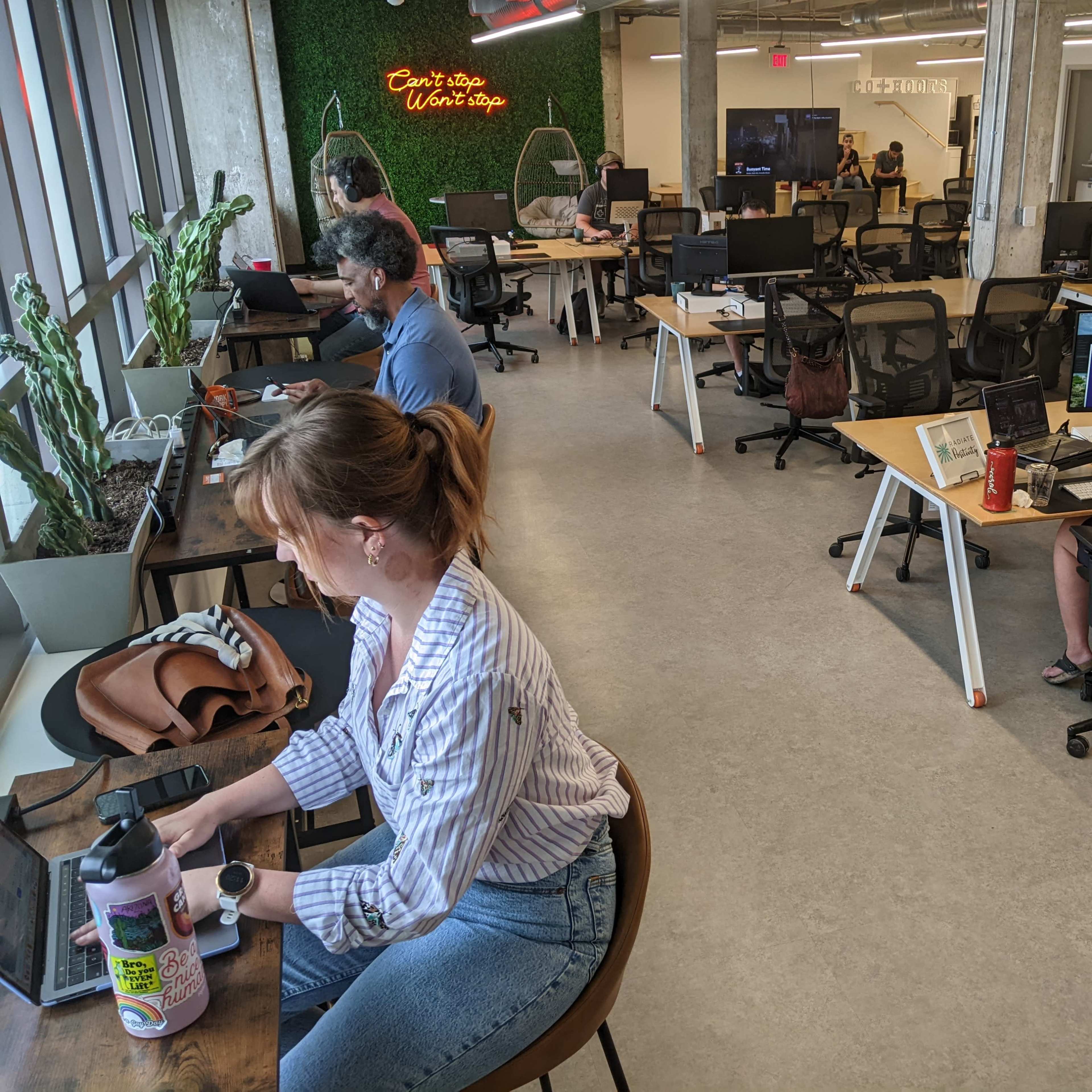 A modern office space features several people working at desks with computers, surrounded by greenery and large windows.