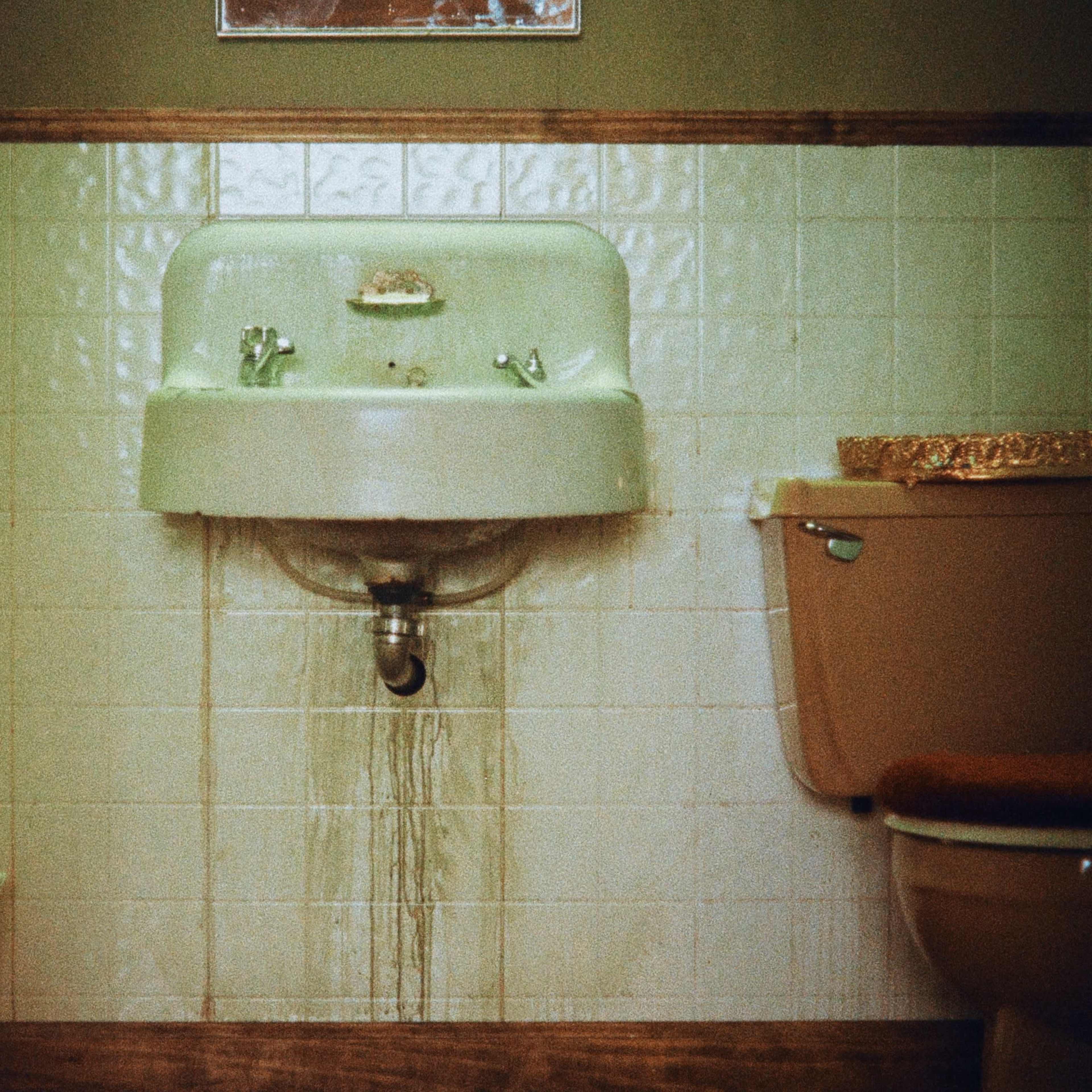 A bathroom with a green sink and a toilet, featuring leaking water from the sink onto the tiled floor.