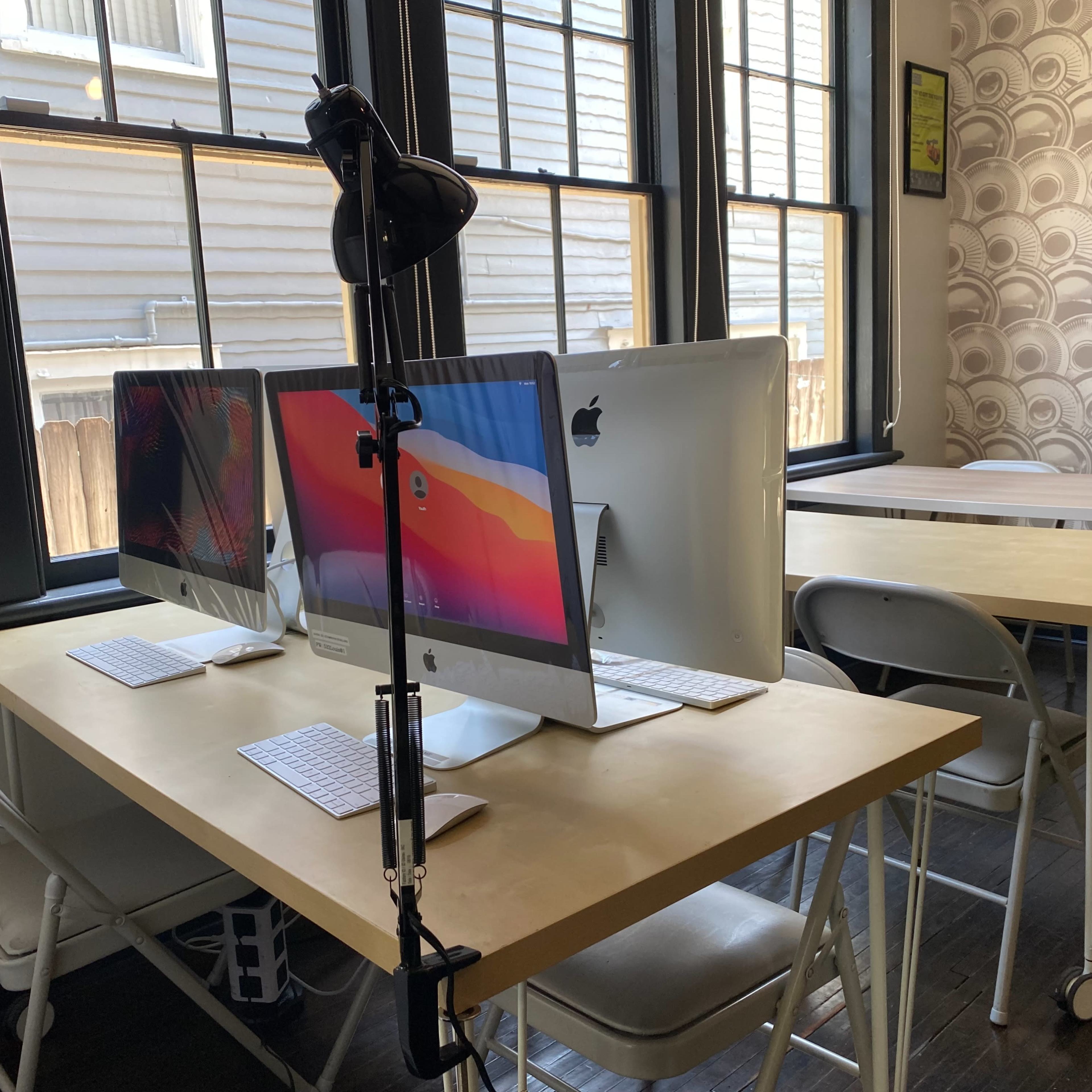 A workspace features two iMac computers on a table, accompanied by folding chairs and a lighting stand.