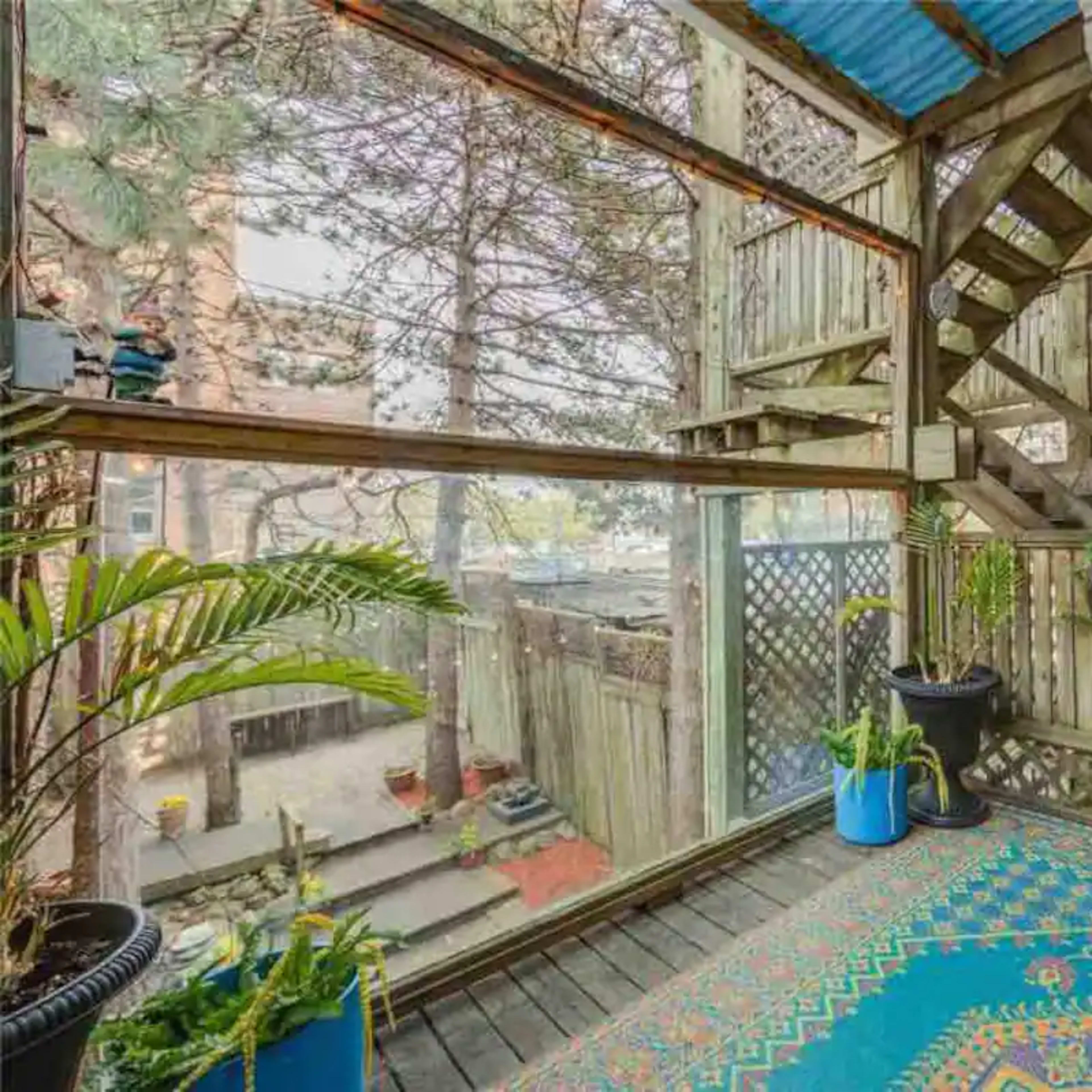 A wooden deck area with large glass panels, a blue patterned rug, potted plants, and a staircase leading to an upper level.