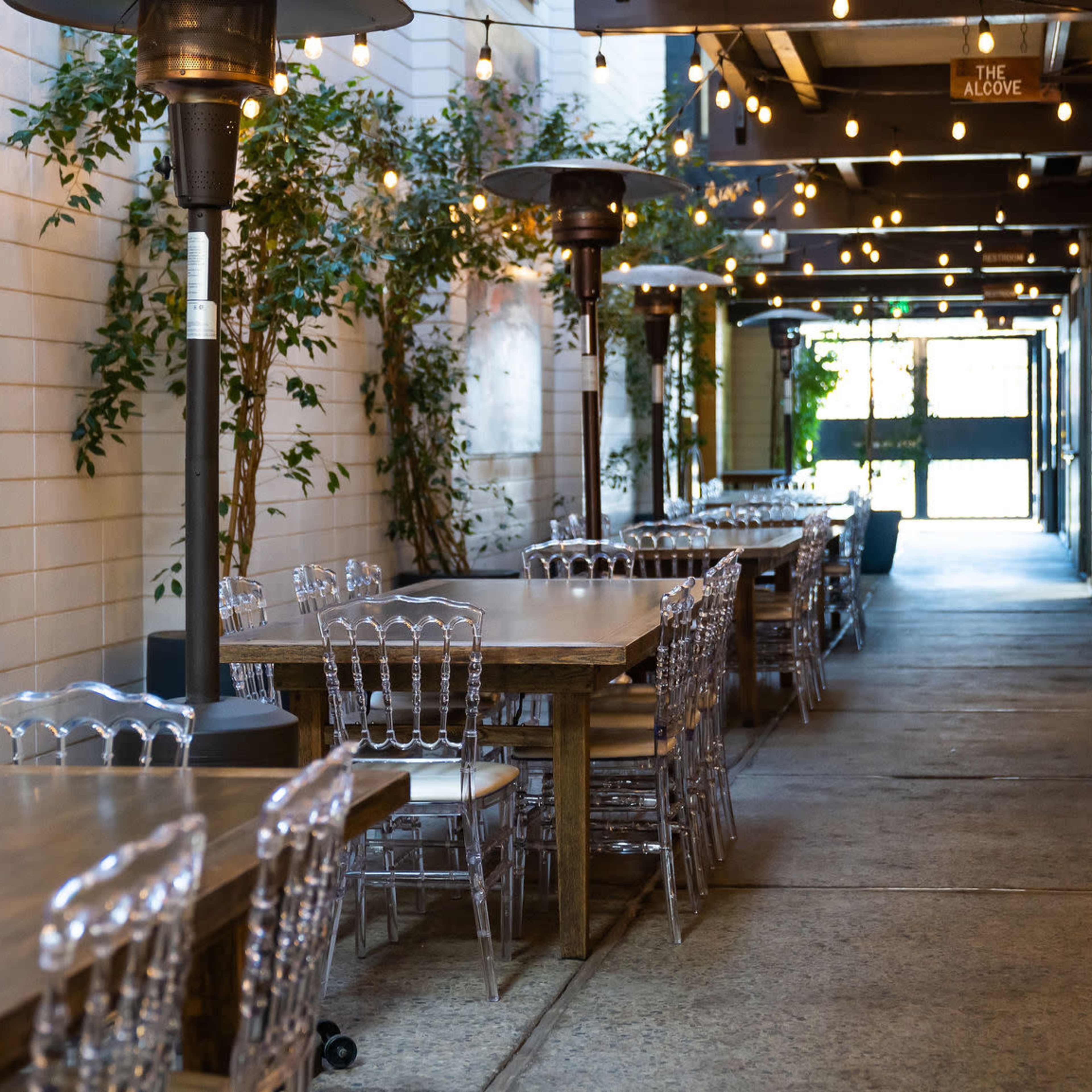 A narrow outdoor dining area lined with tables and illuminated by string lights.