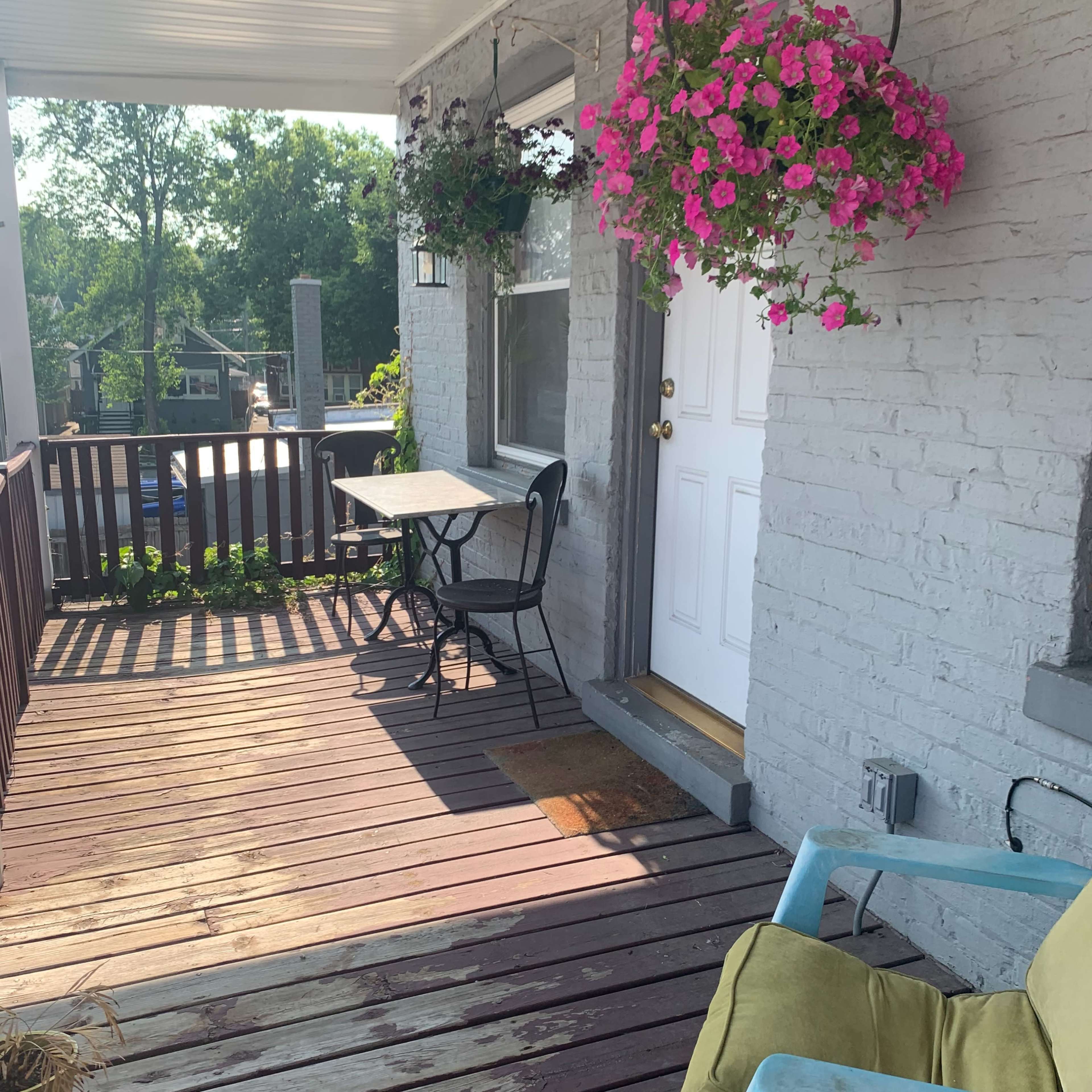 A small porch features a table and chair, potted plants, and hanging flower baskets.