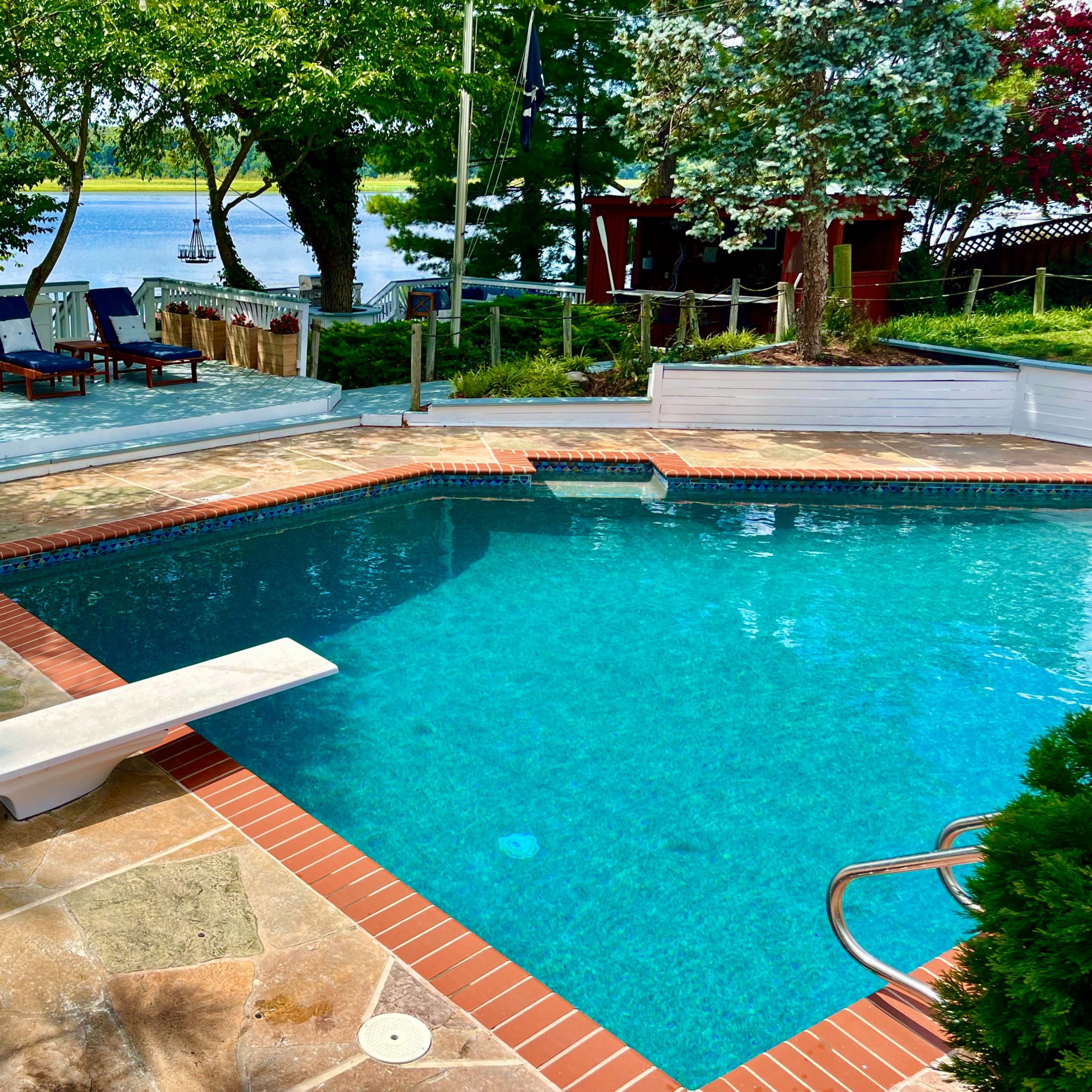 A swimming pool with a diving board is surrounded by a patio area that includes lounge chairs and overlooks a lake.
