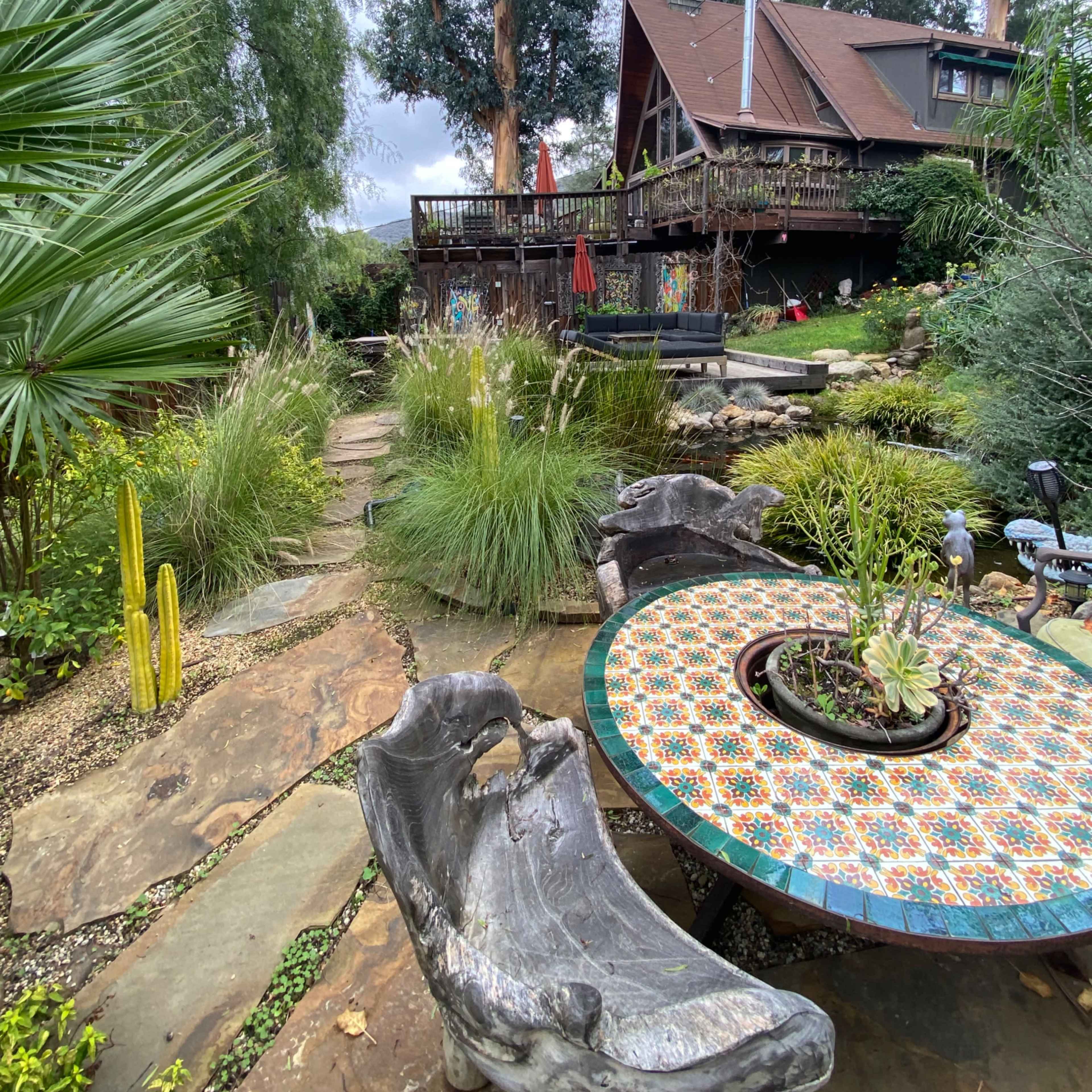 A stone pathway leads through a lush garden to a large wooden deck and a rustic house surrounded by tall trees.