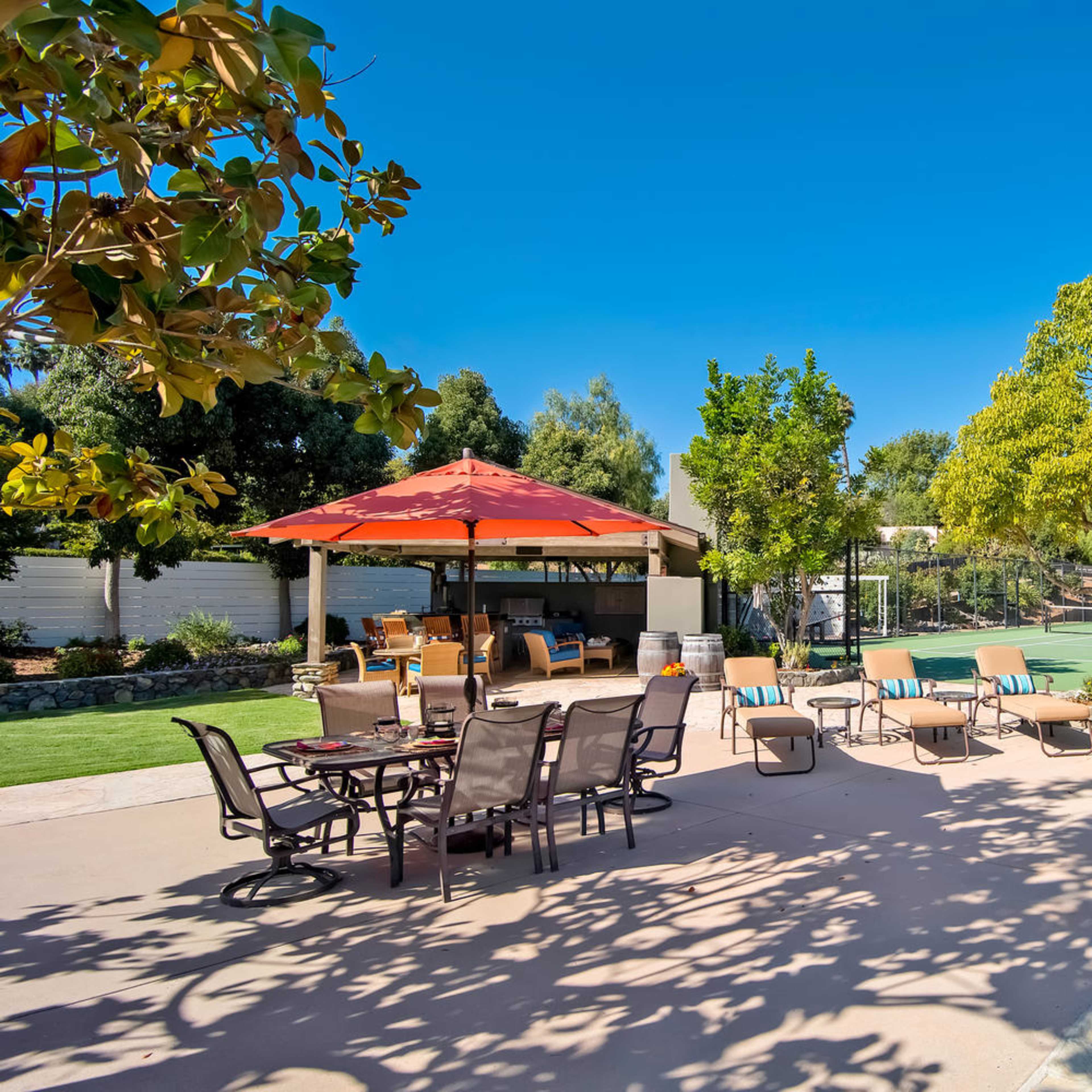 A sunny backyard area with seating around a shaded table and a tennis court in the background.