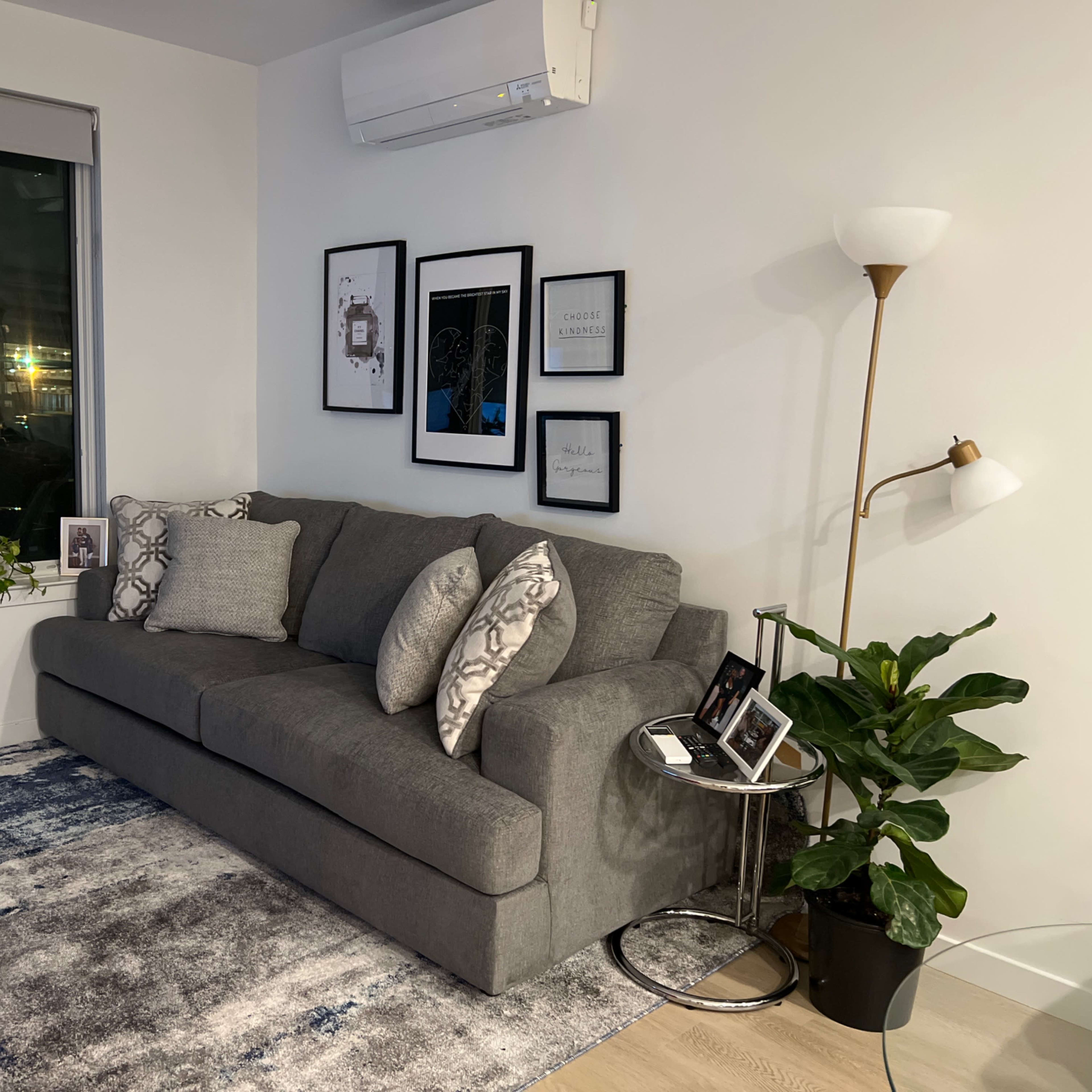 A gray sectional sofa is positioned against a white wall, surrounded by framed artwork and a tall plant, with a lamp and decorative items on a side table.