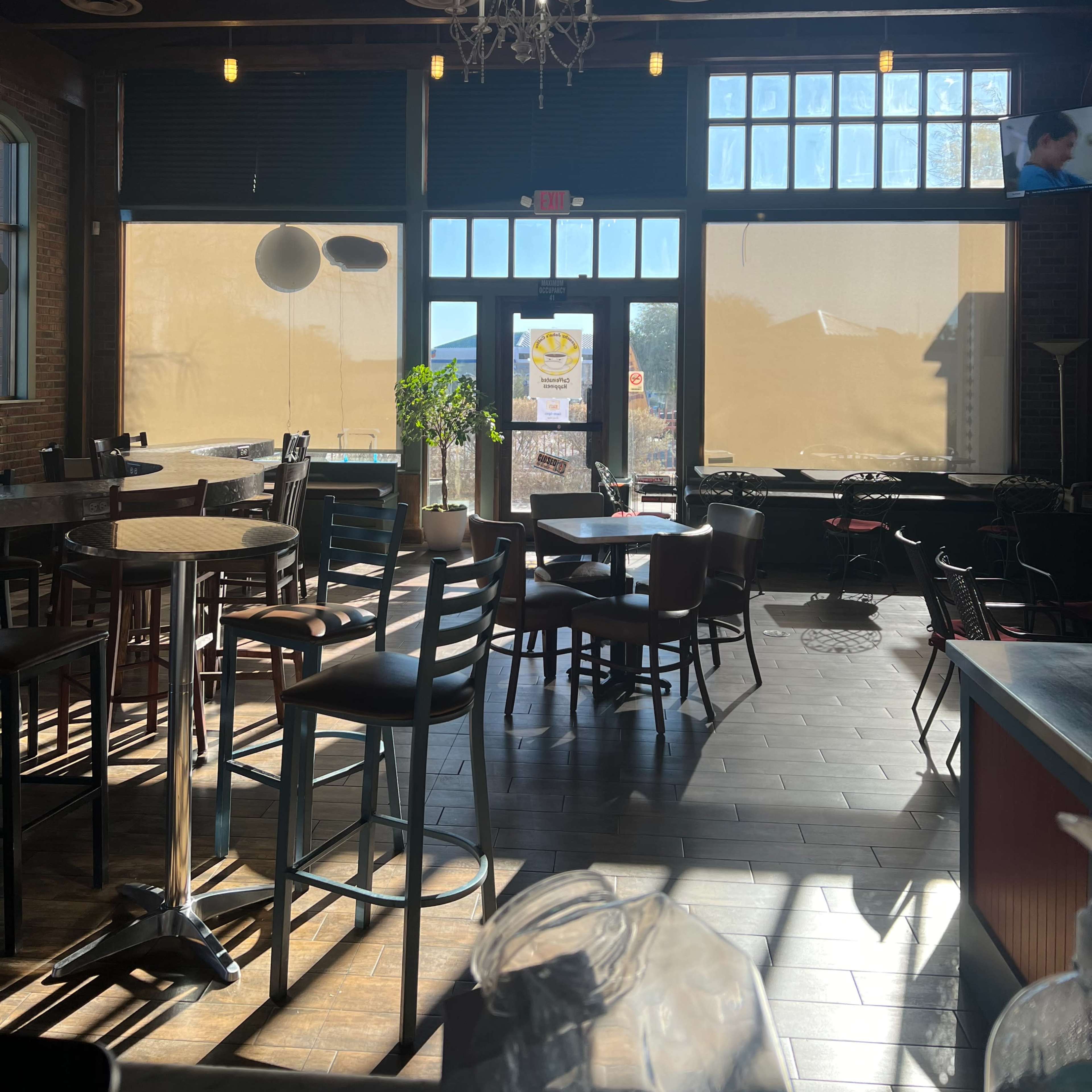 The interior of a restaurant with wooden furniture, large windows covered partially with shades, and hanging light fixtures.