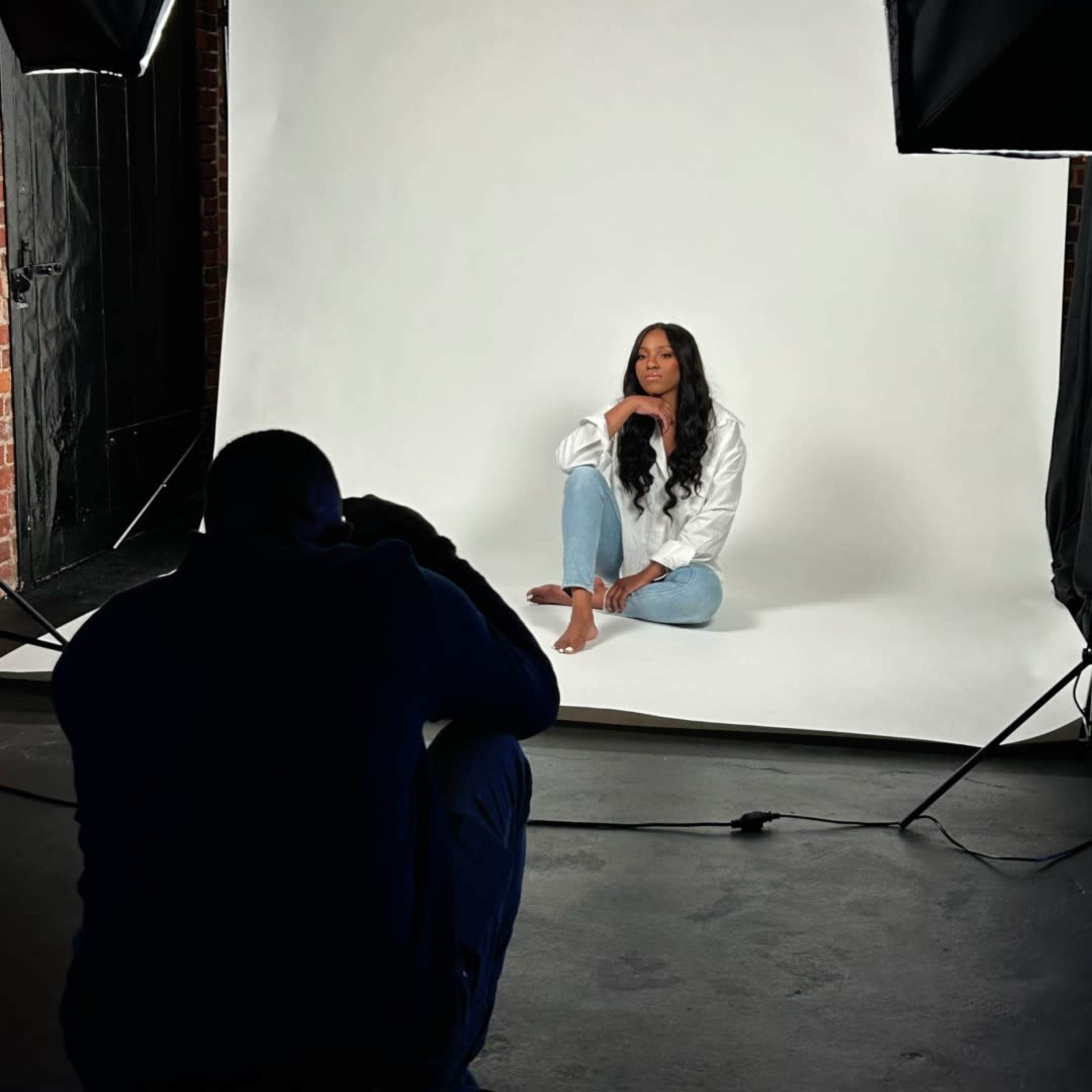 A photographer captures a seated model in a white studio with a plain backdrop.