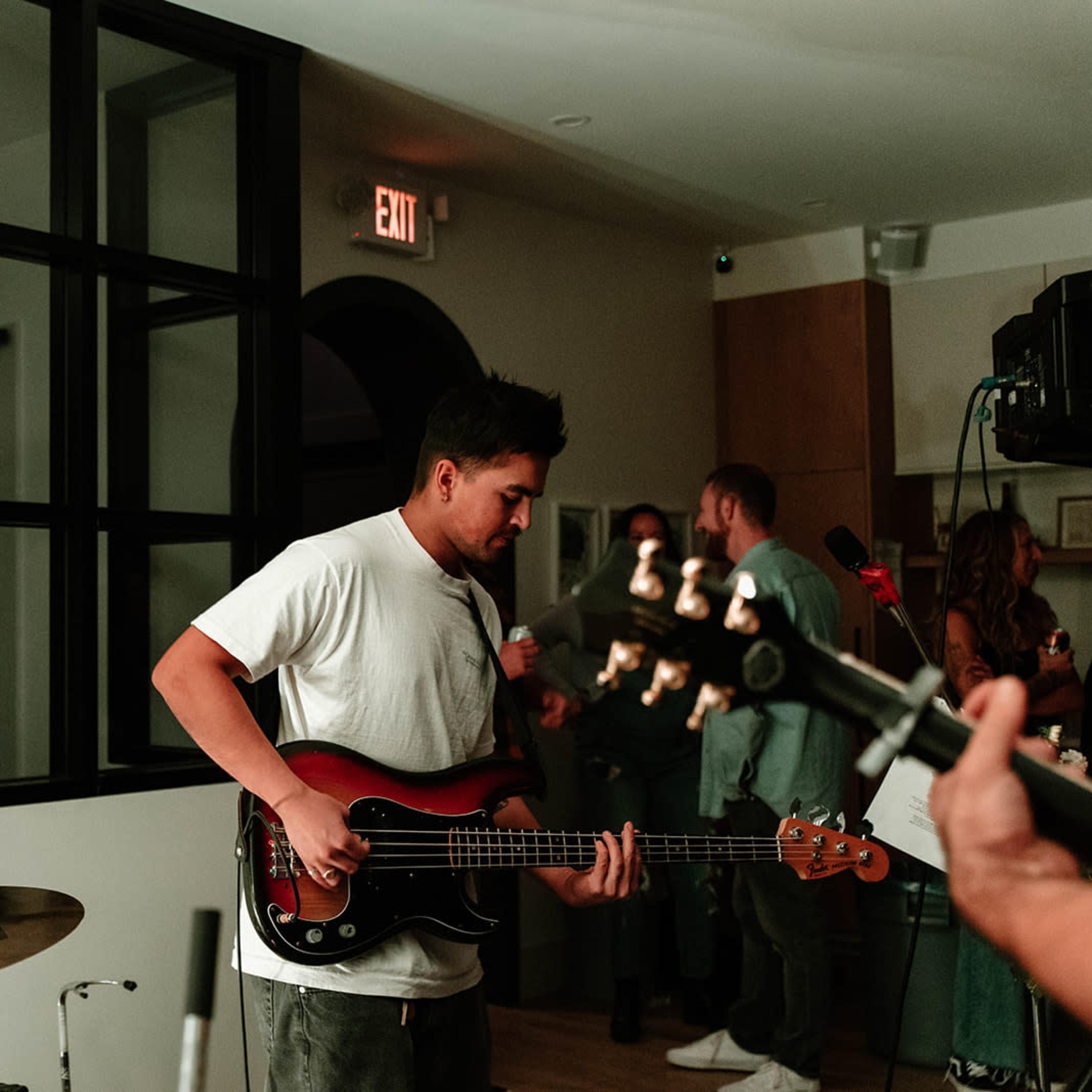 A musician plays a bass guitar in a dimly lit room with large windows and a small crowd in the background.