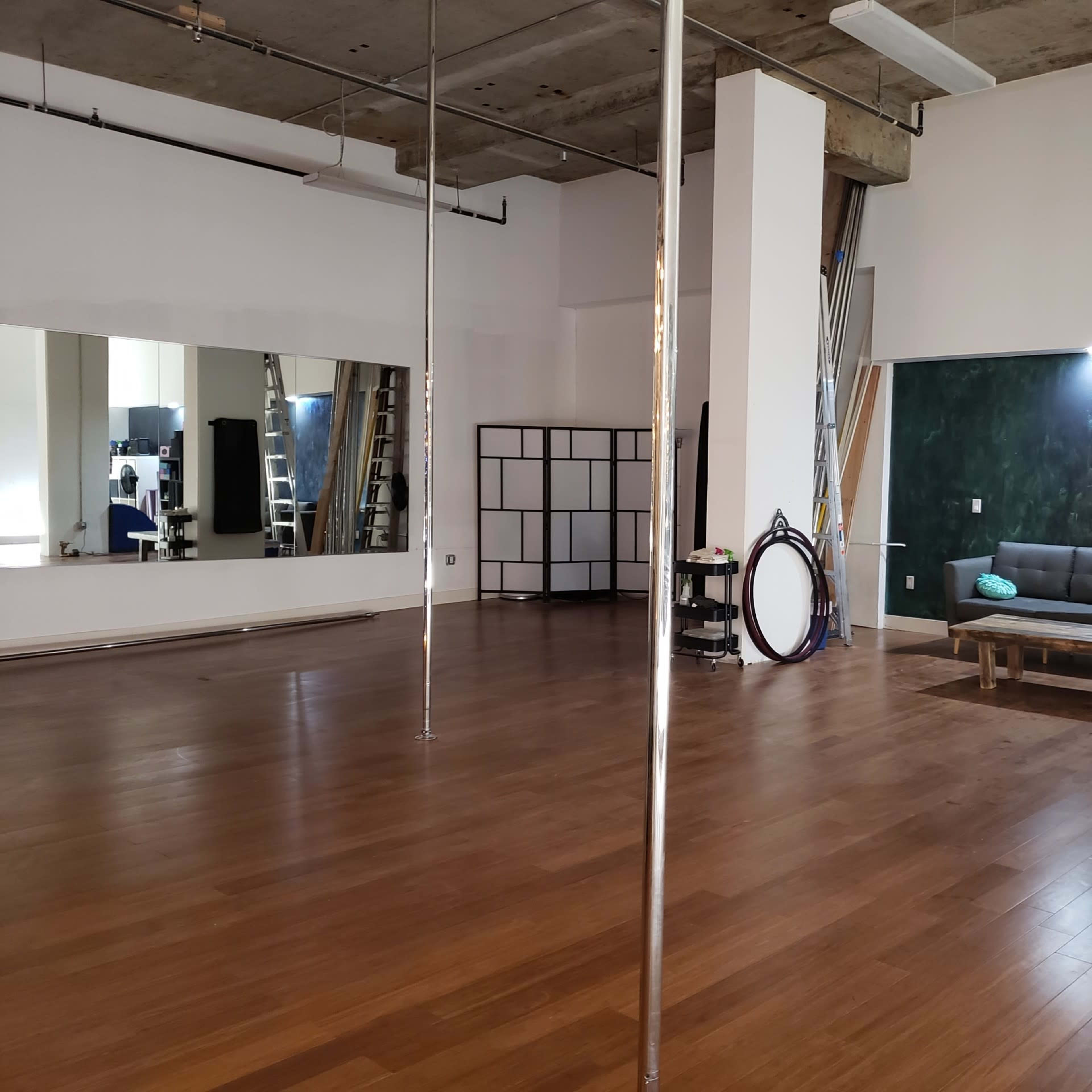 A spacious studio with wooden flooring, featuring several pole dance apparatuses, a mirror on one wall, and a sofa set against a green accent wall.
