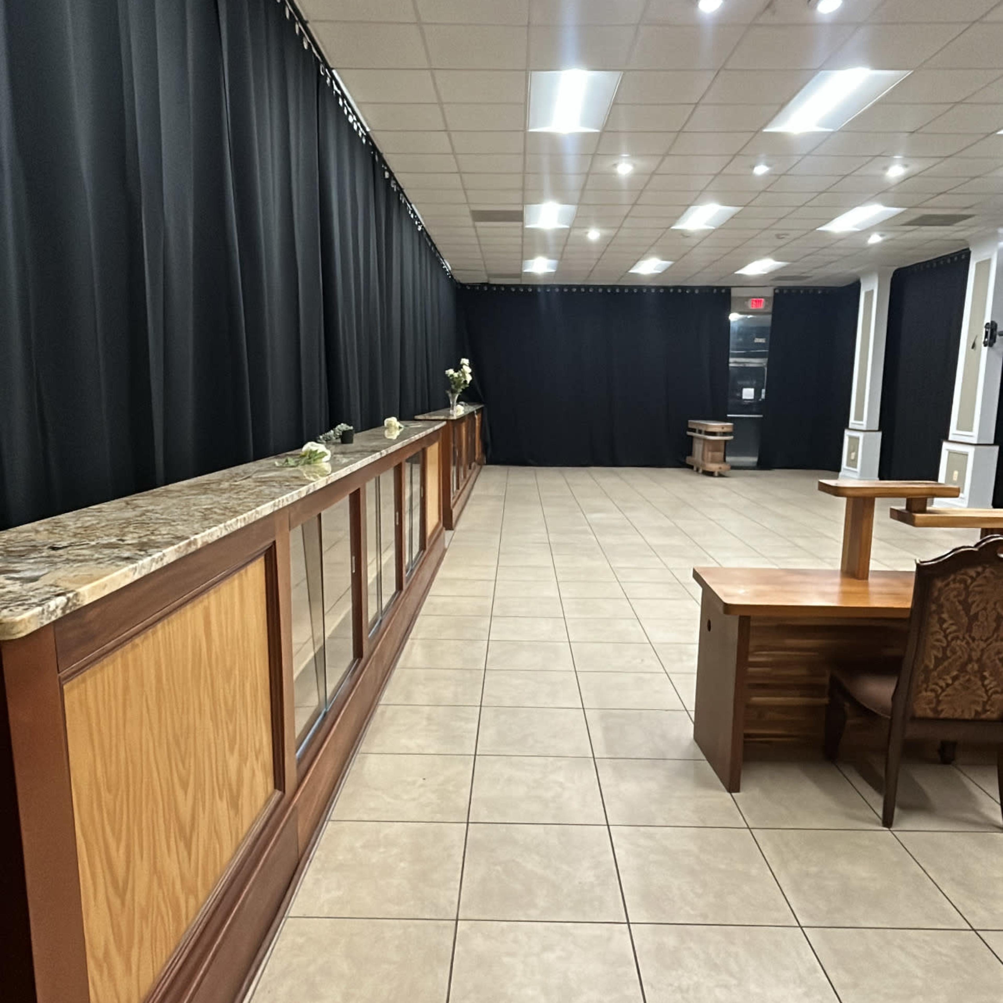 A spacious room with tiled flooring, a long wooden counter against one wall, and a table in the center, surrounded by black curtains.