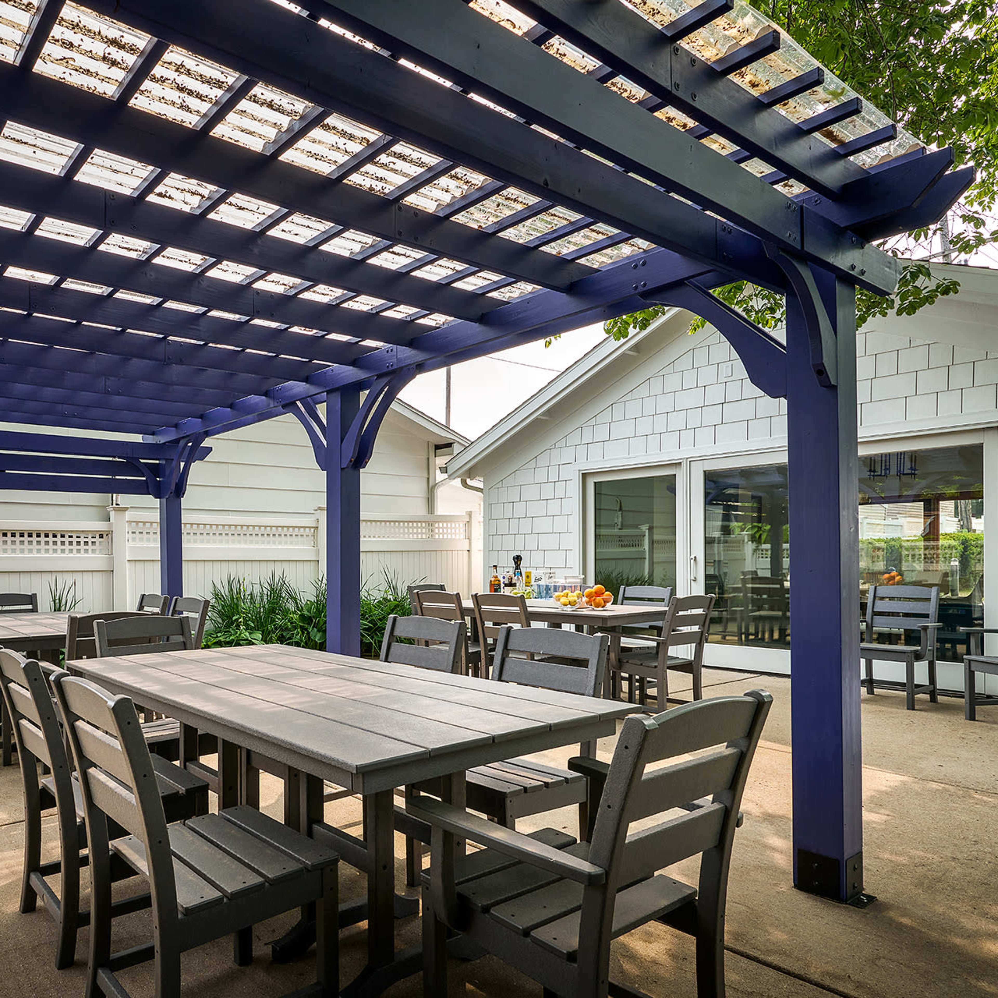 A spacious outdoor dining area with a purple pergola, wooden tables and chairs, and a house in the background.