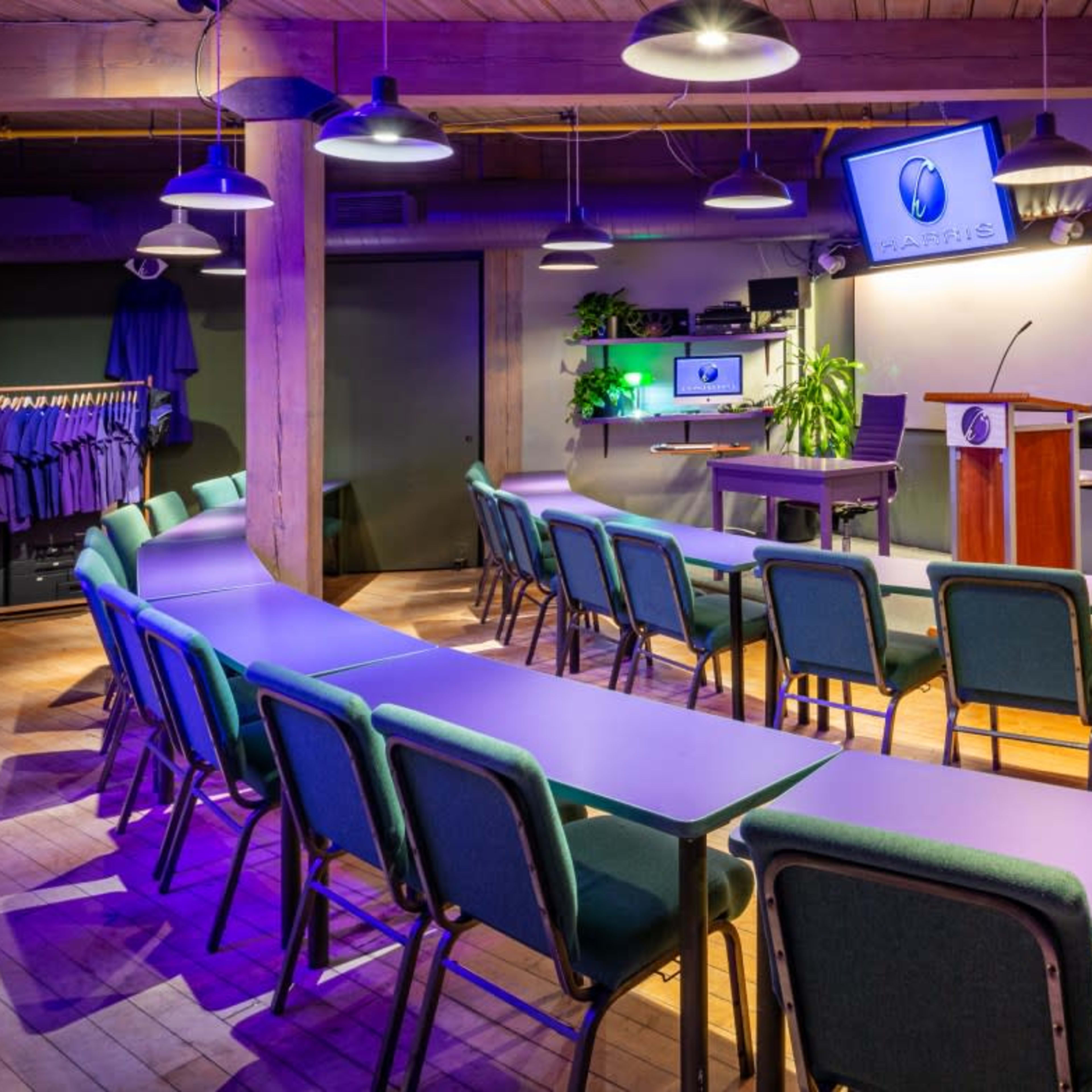 A classroom or meeting space with rows of tables and chairs arranged facing a podium and screens, surrounded by plants and hanging lights.
