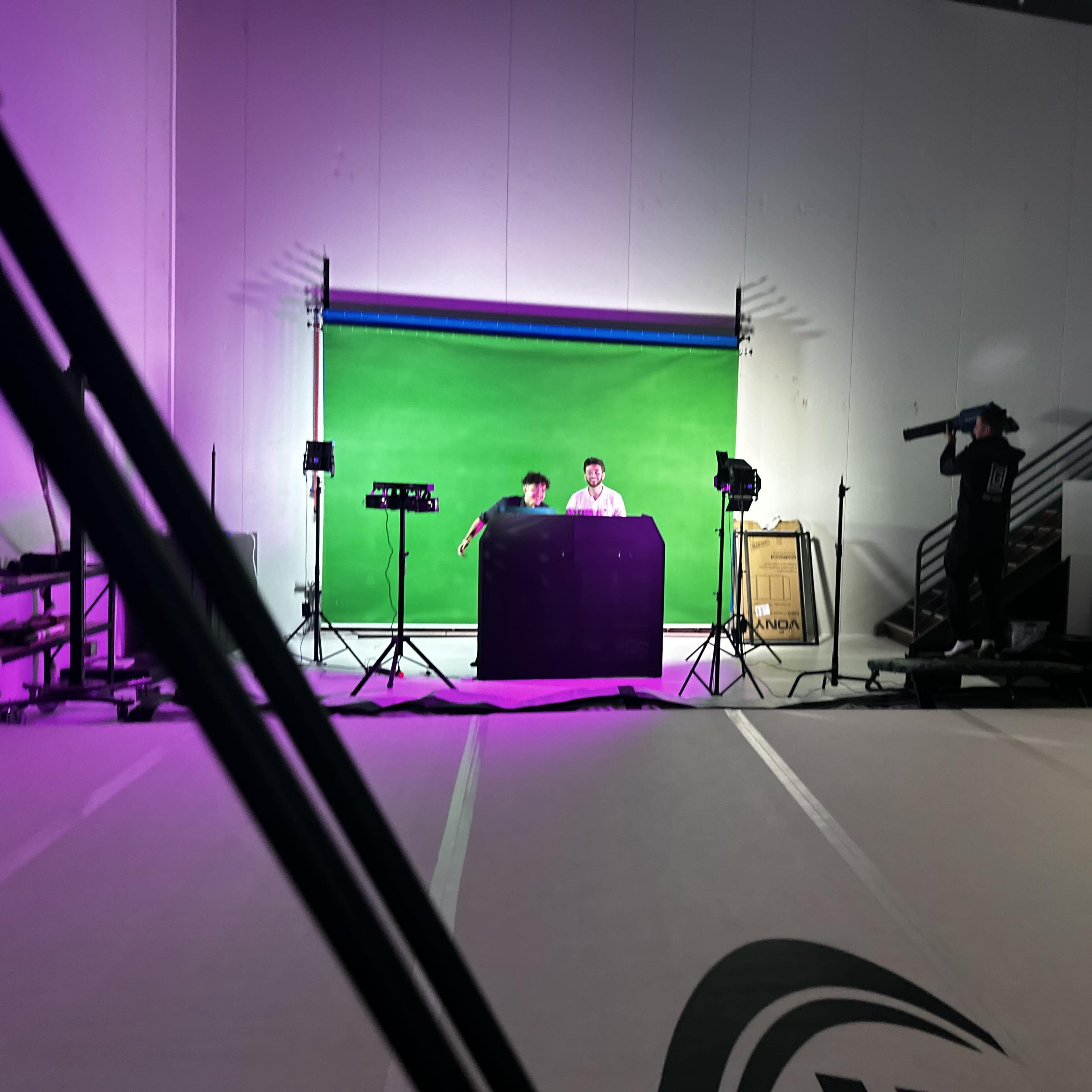 A green screen setup is seen in a studio, where two individuals are positioned at a table while another person stands nearby with a camera.