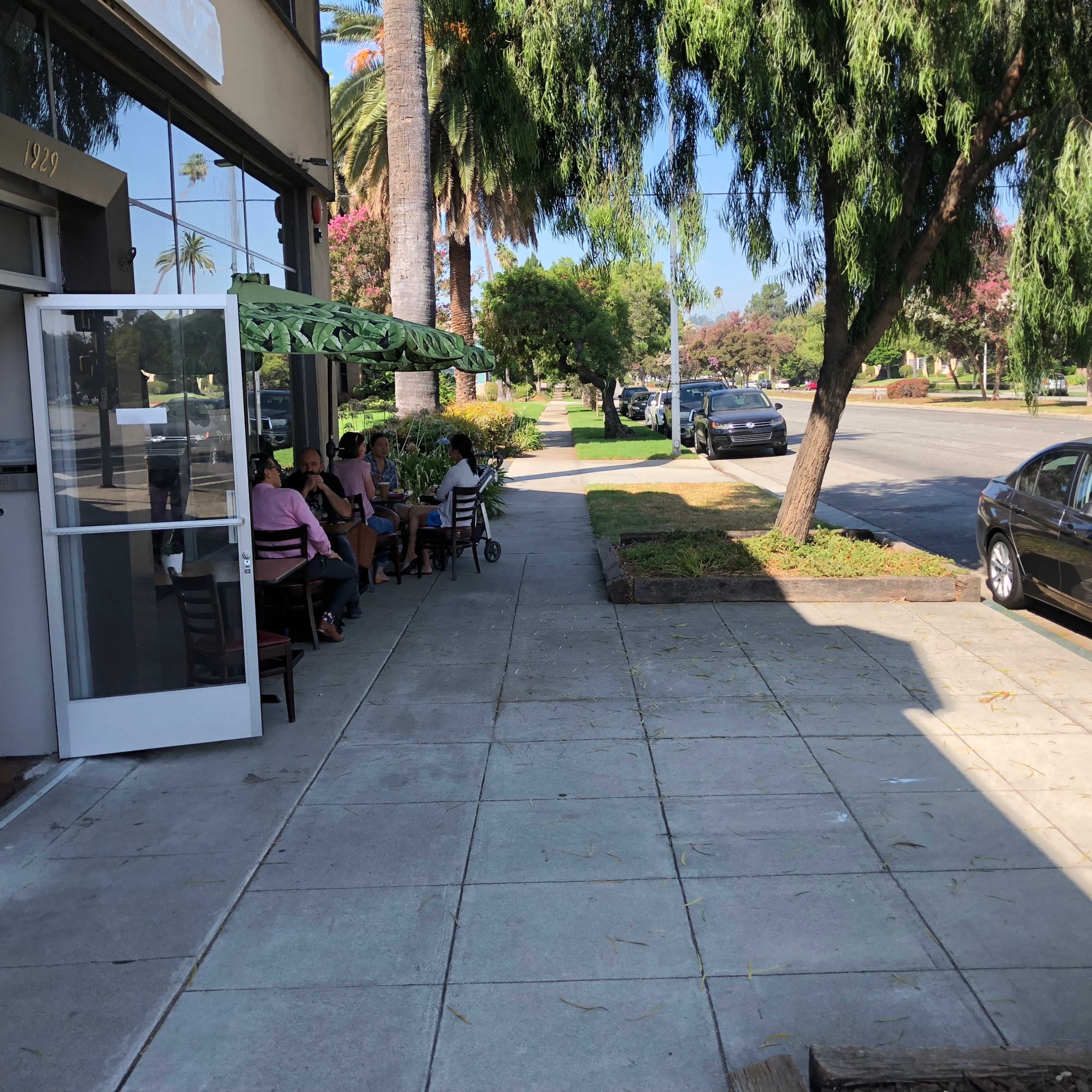 A sidewalk runs alongside a café with outdoor seating on one side and a street lined with parked cars on the other.