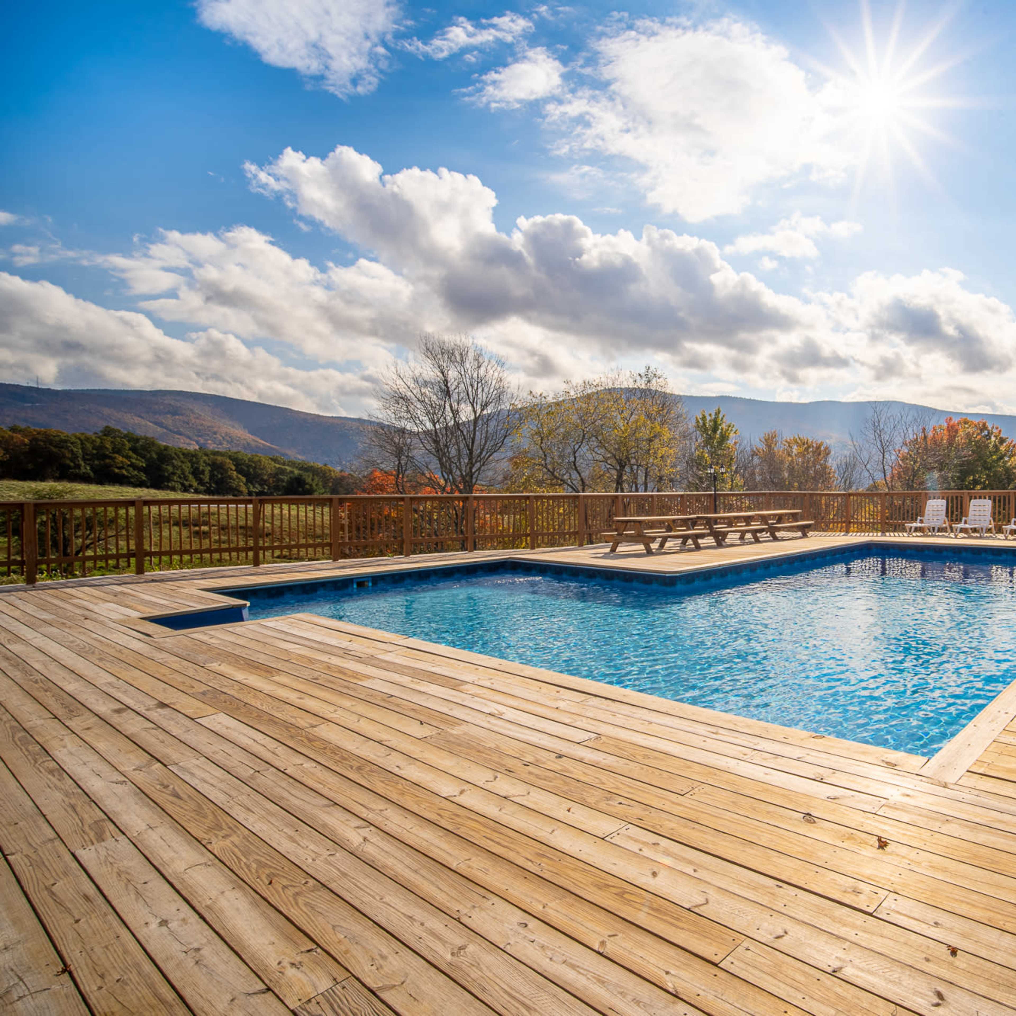 A clear blue swimming pool surrounded by wooden decking and lounge chairs, with mountains and a partly cloudy sky in the background.