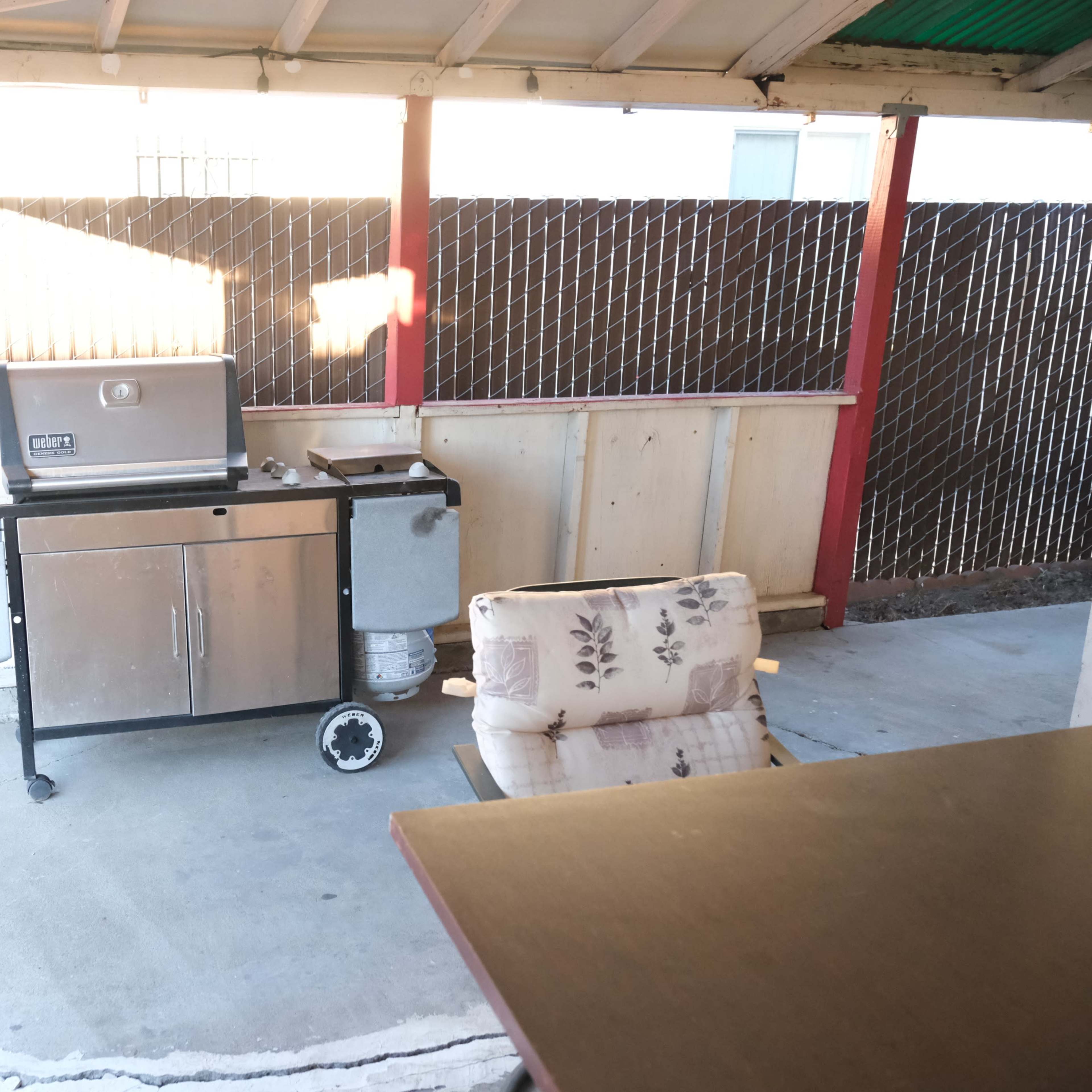 An outdoor patio with a barbecue grill on a cart next to a cushioned chair and a fenced area.