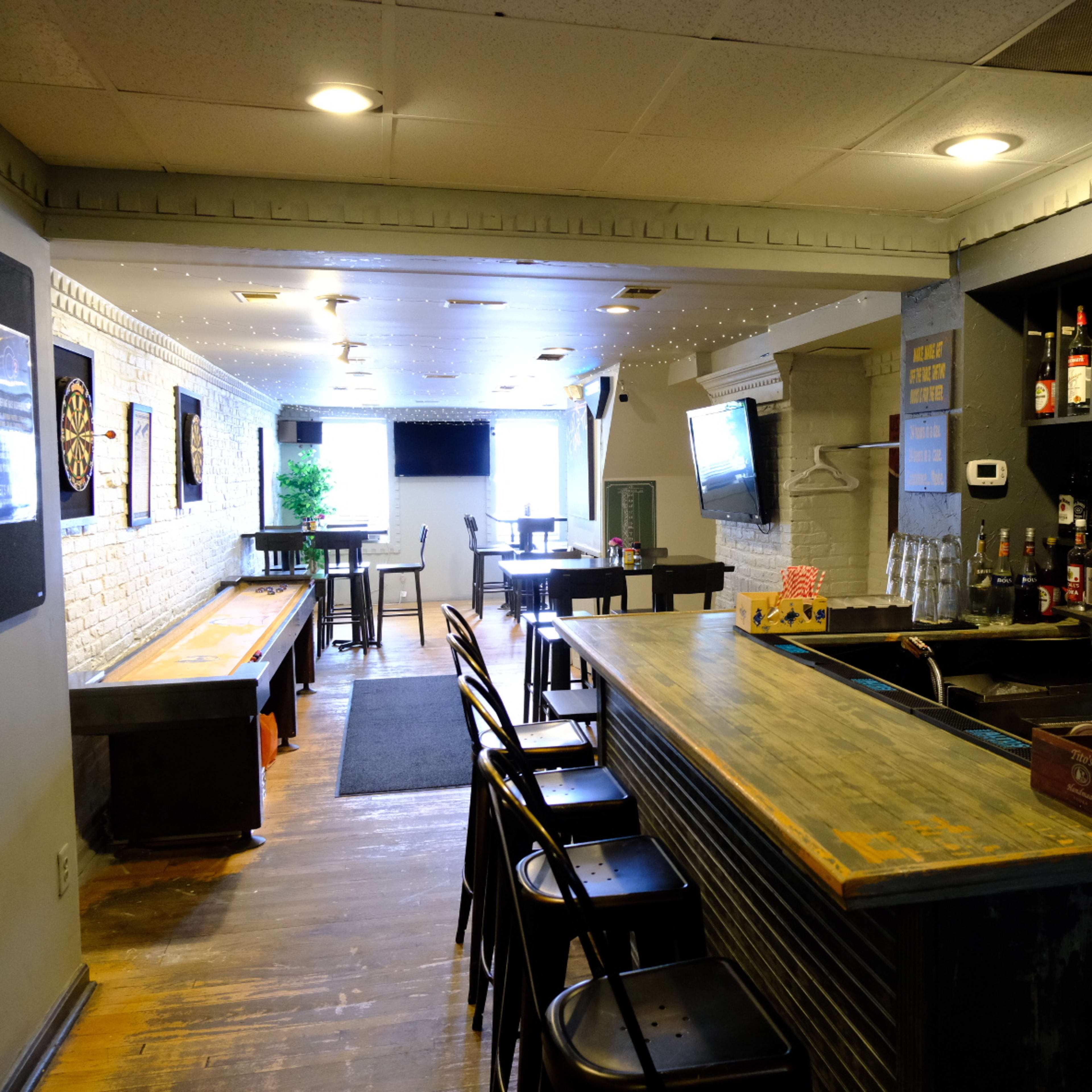 A bar area with a long wooden counter and a shelf stocked with various bottles, leading to a wider seating area with tables and a dartboard setup.