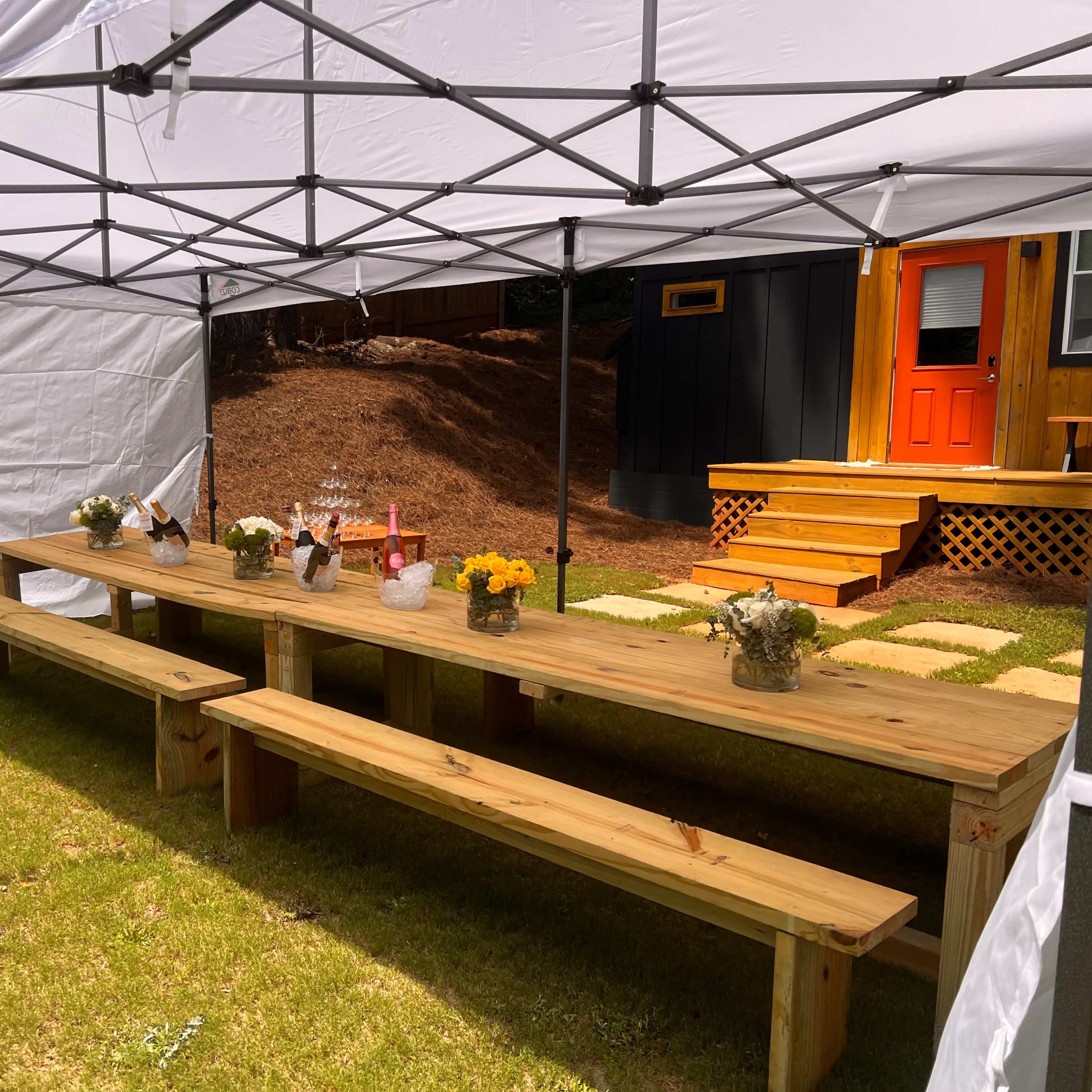 A wooden table with several vases of flowers is set up under a white canopy in a grassy area, leading to a house with an orange door.