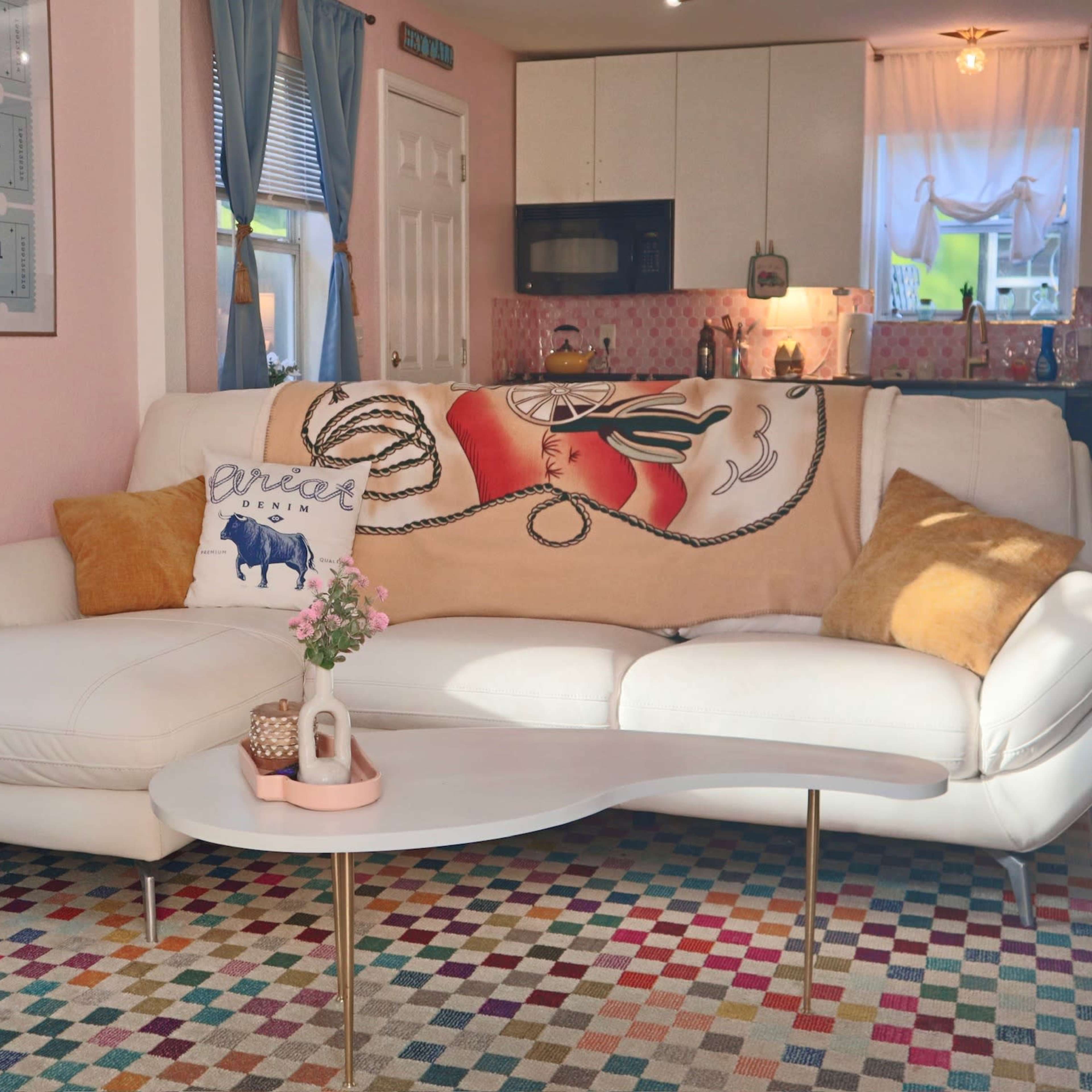 A modern living room features a white sectional sofa with colorful throw pillows, a patterned rug, and a coffee table in front of a kitchen area with pastel-colored decor.