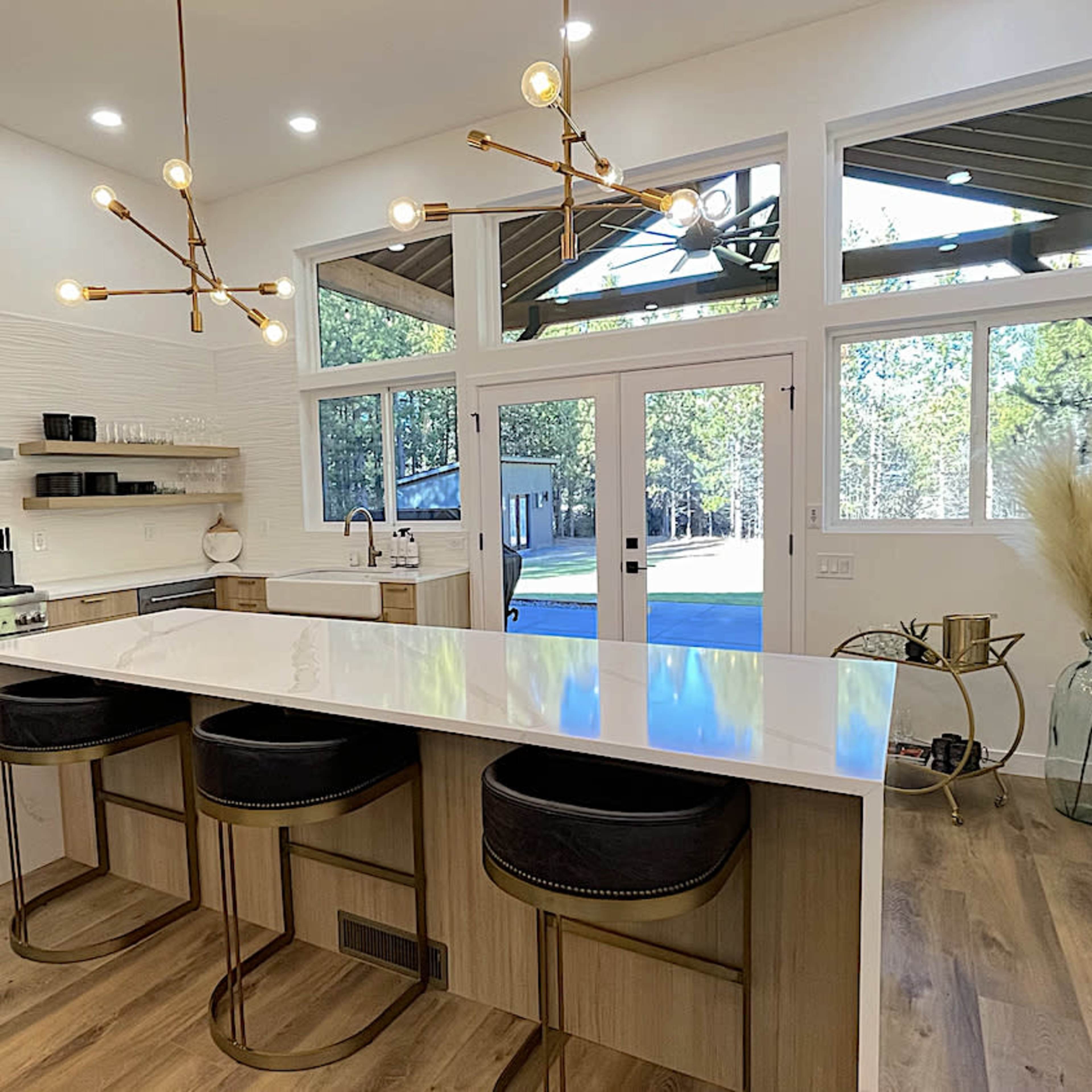 A modern kitchen with a large island fitted with black stools, stainless steel appliances, and large windows overlooking a wooded area.