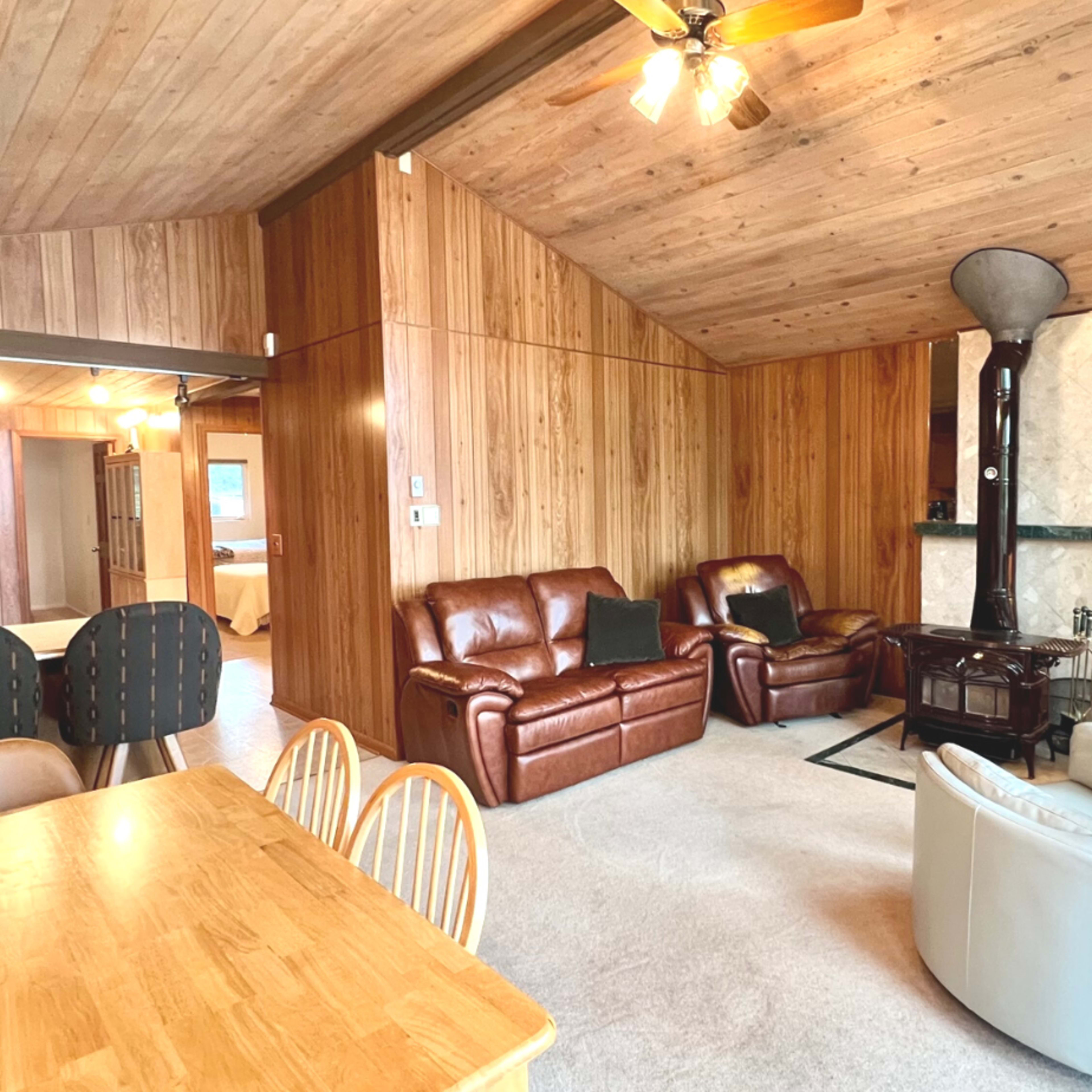 A cozy living area featuring wooden walls, a dining table with chairs, leather sofas, and a corner fireplace.