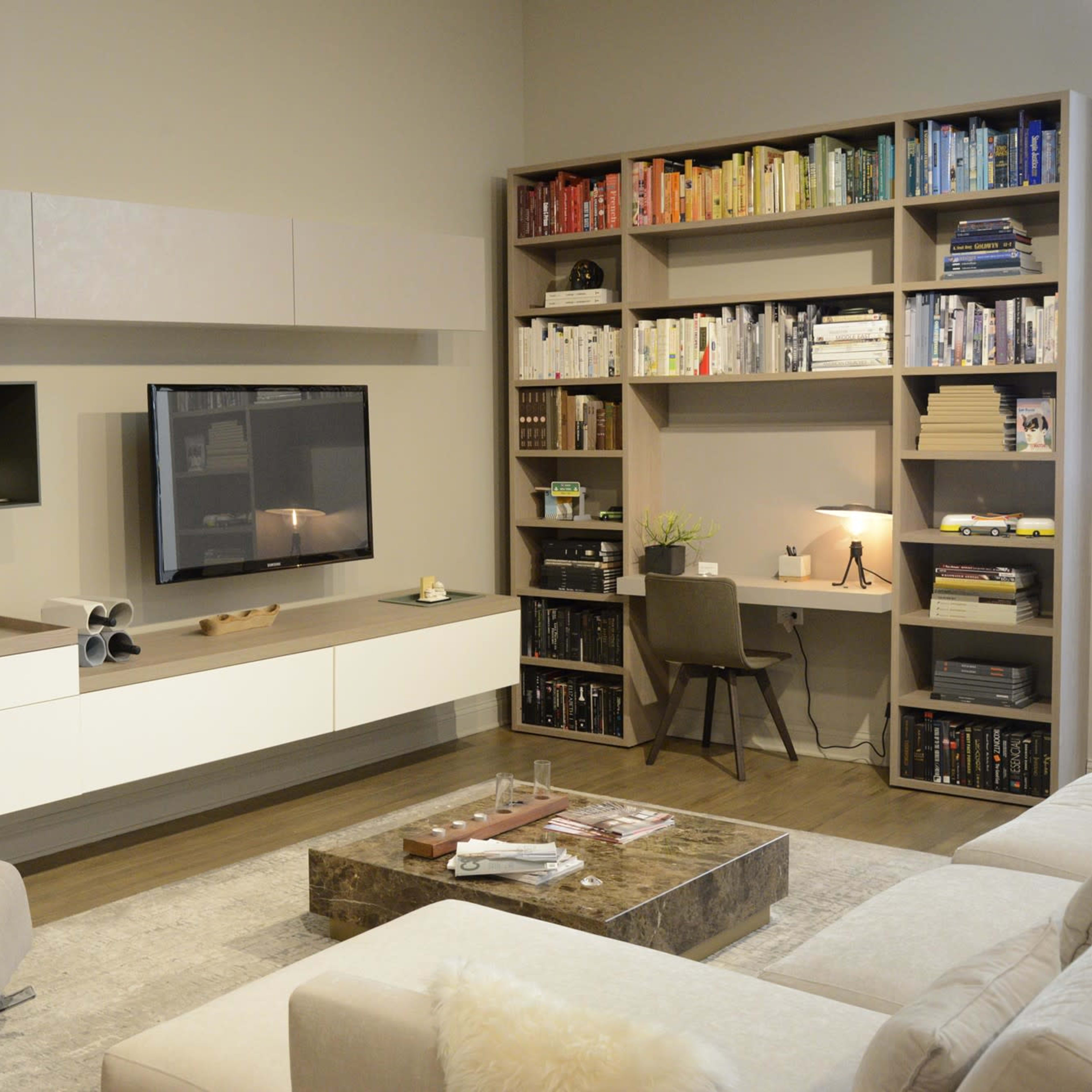 A modern living room features a light-colored wall unit with a TV, bookshelves filled with books, a cozy armchair, and a coffee table on a rug.