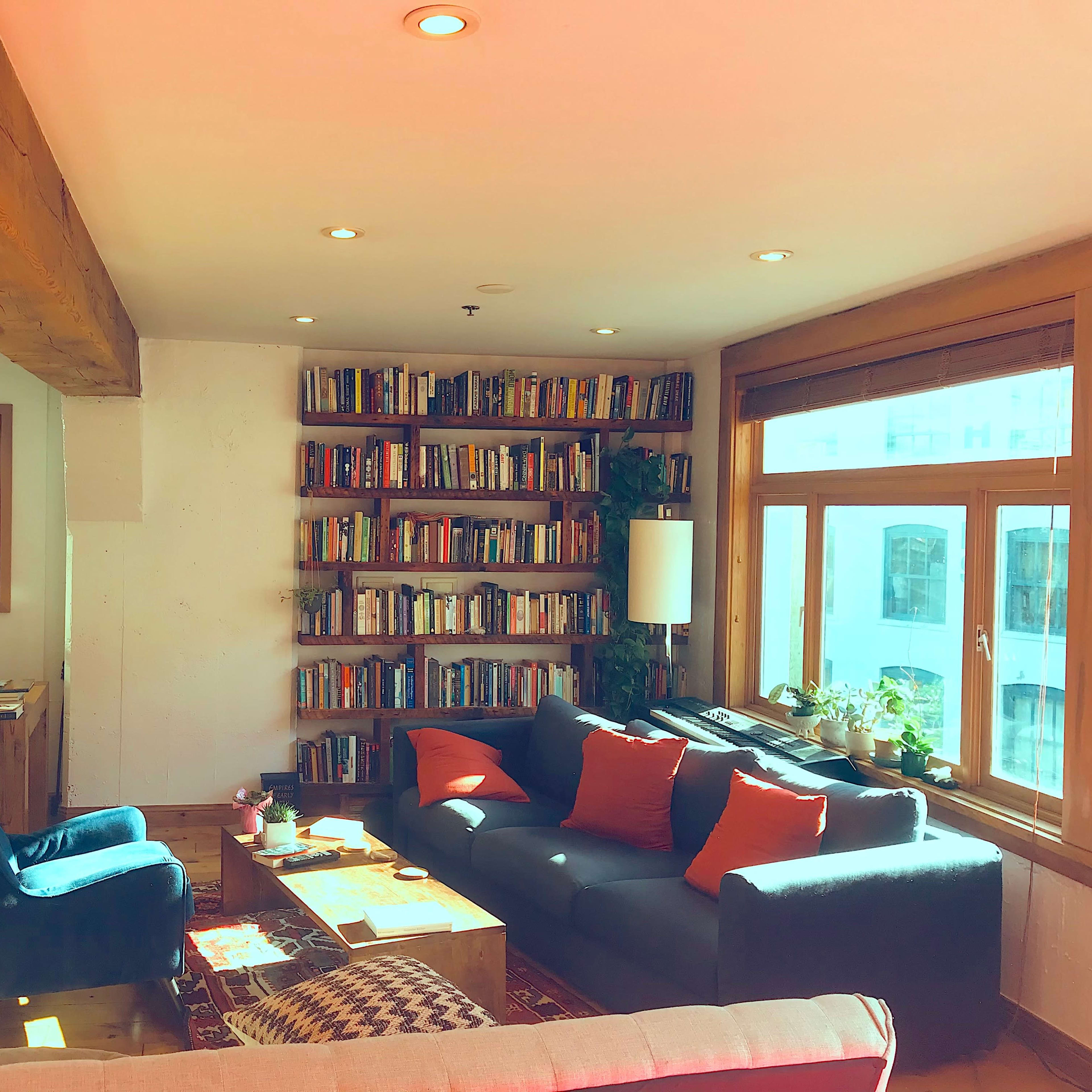 A cozy living room with a bookshelf filled with books, a sofa, and large windows providing natural light.