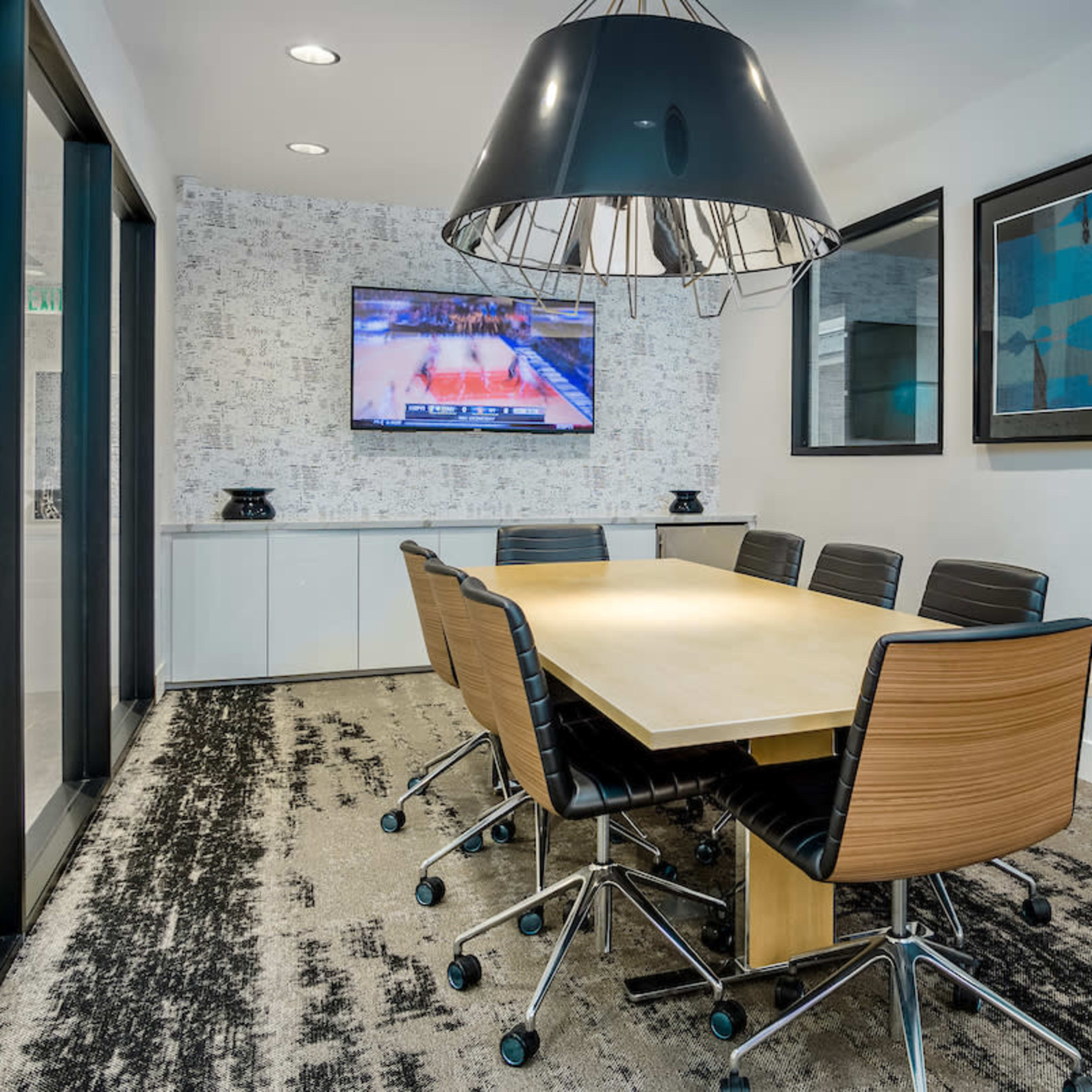A modern conference room features a large wooden table surrounded by black chairs, with a television displaying a basketball game on the wall.