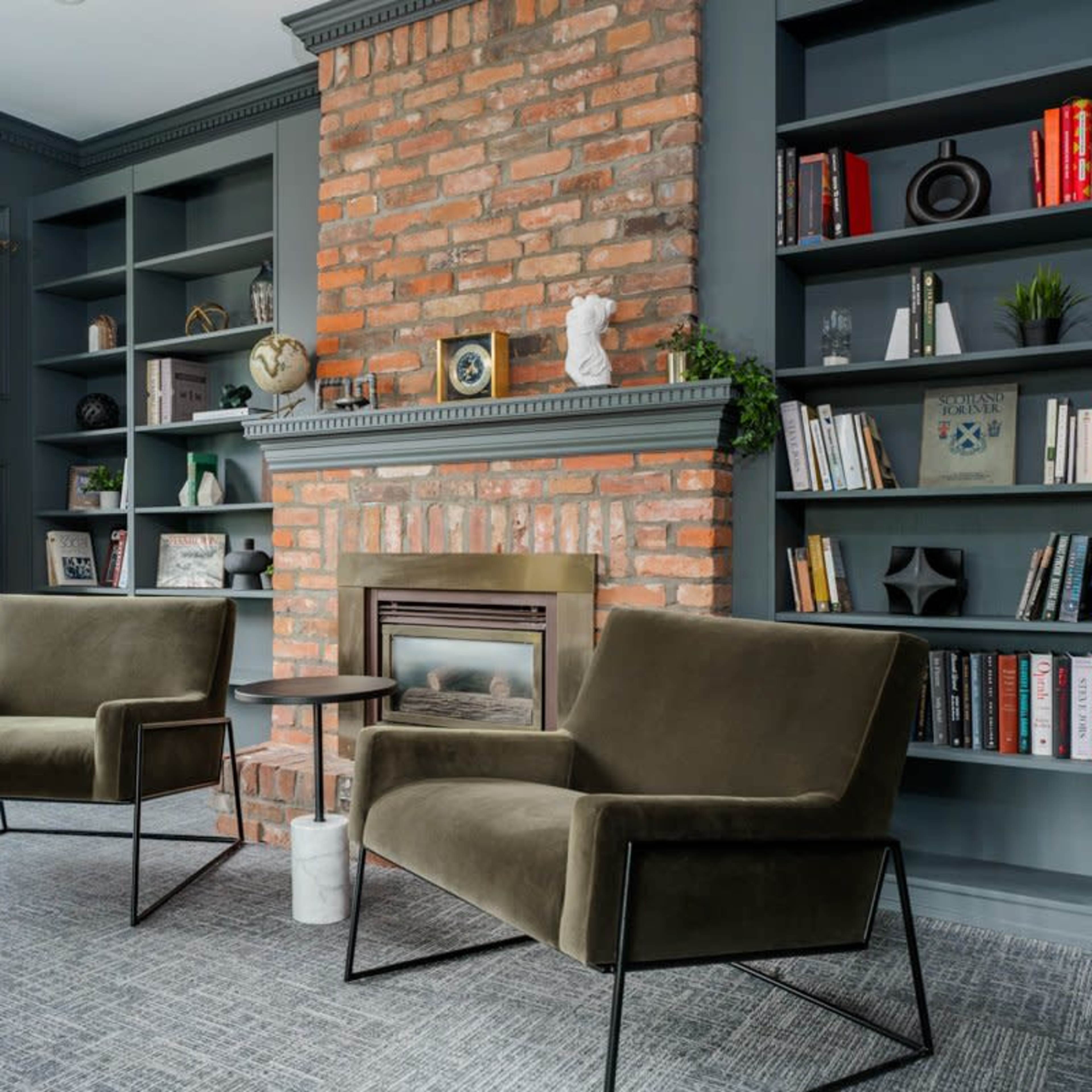 A cozy reading nook features two green chairs, a small table, and a brick fireplace surrounded by built-in bookshelves filled with books and decorative items.