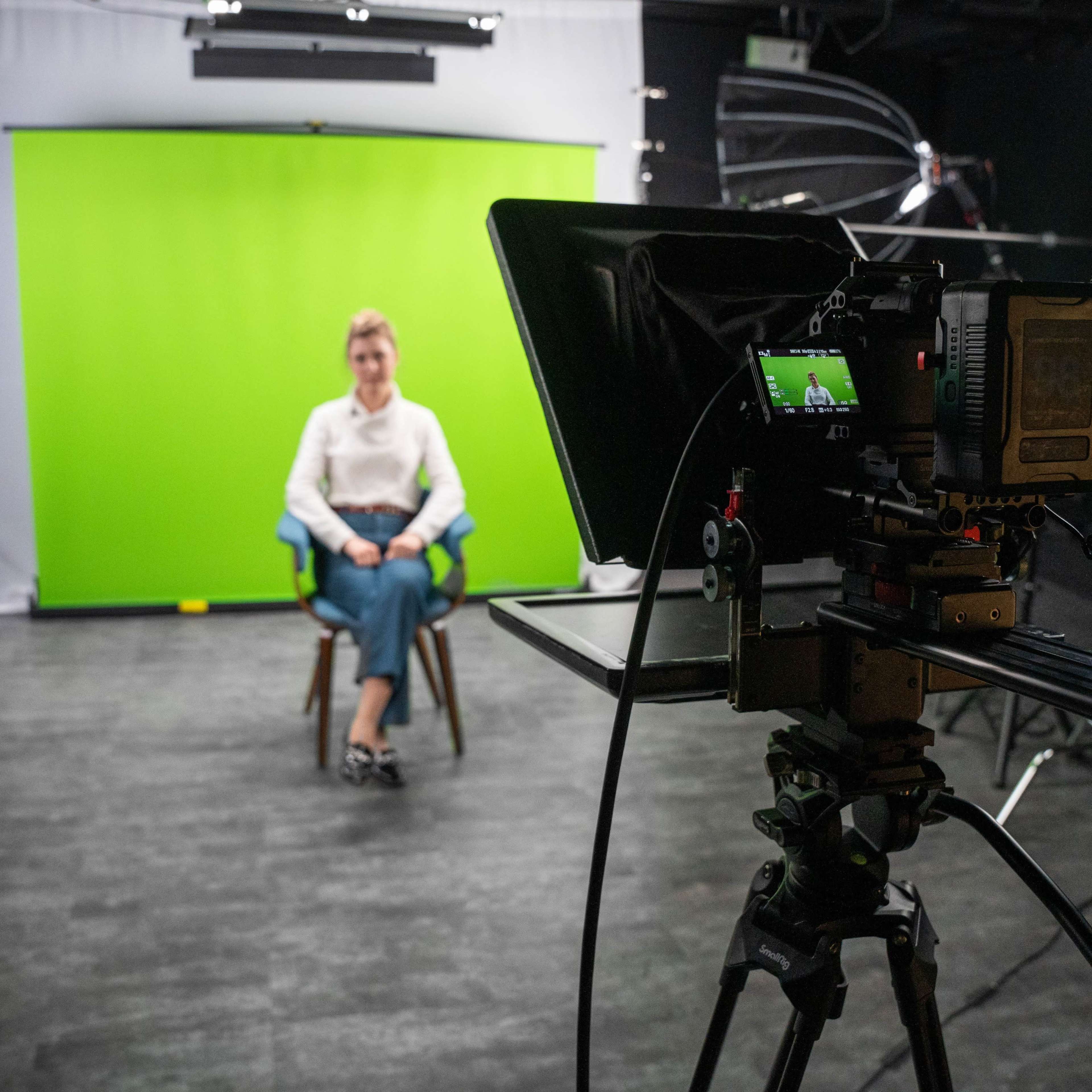 A woman sits in front of a green screen in a photo studio, while a camera on a tripod is focused on her.