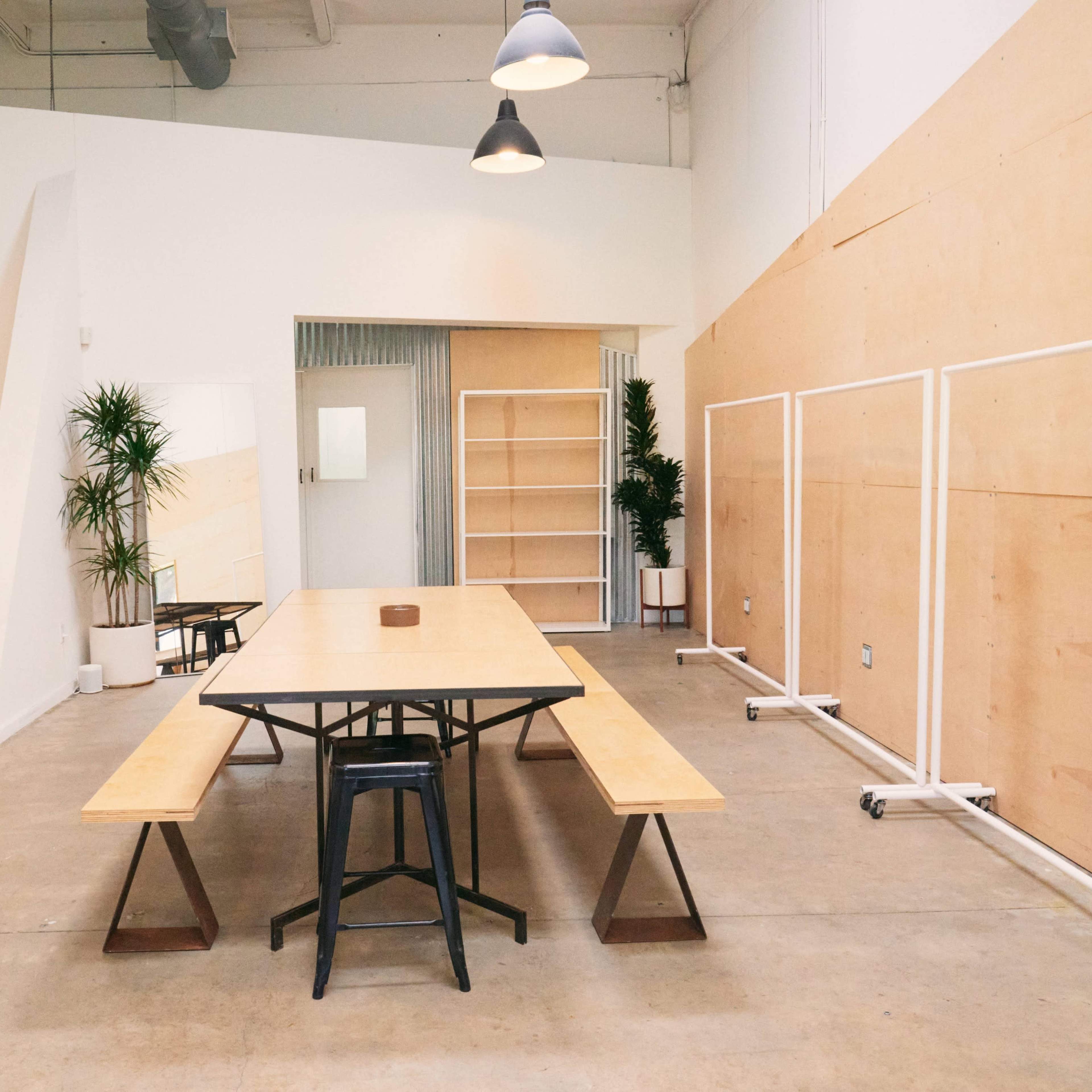 A spacious, modern room with a long wooden table and benches, surrounded by minimalistic decor and plants.