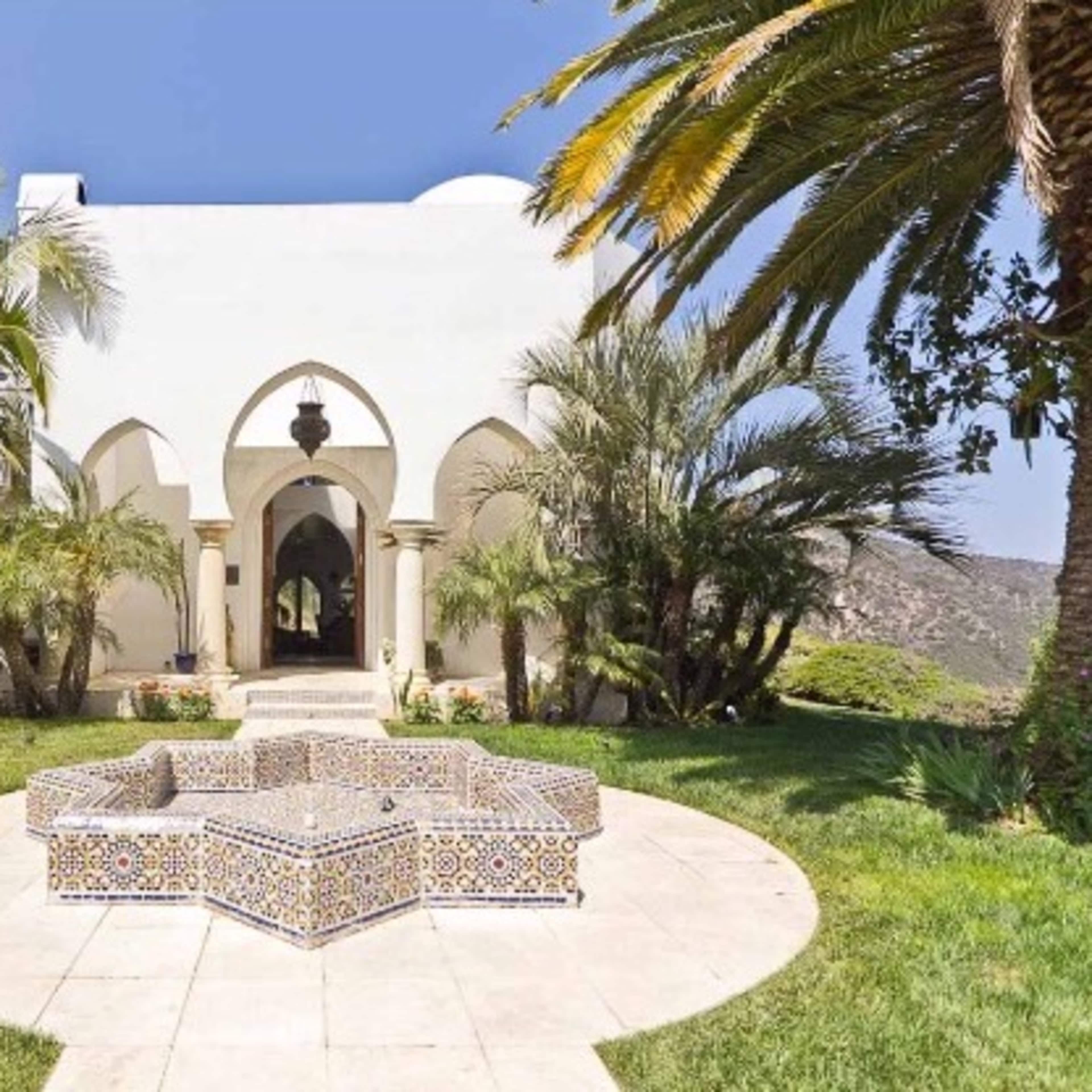 A white villa with arched doorways, surrounded by palm trees and a tiled seating area on a circular stone patio.
