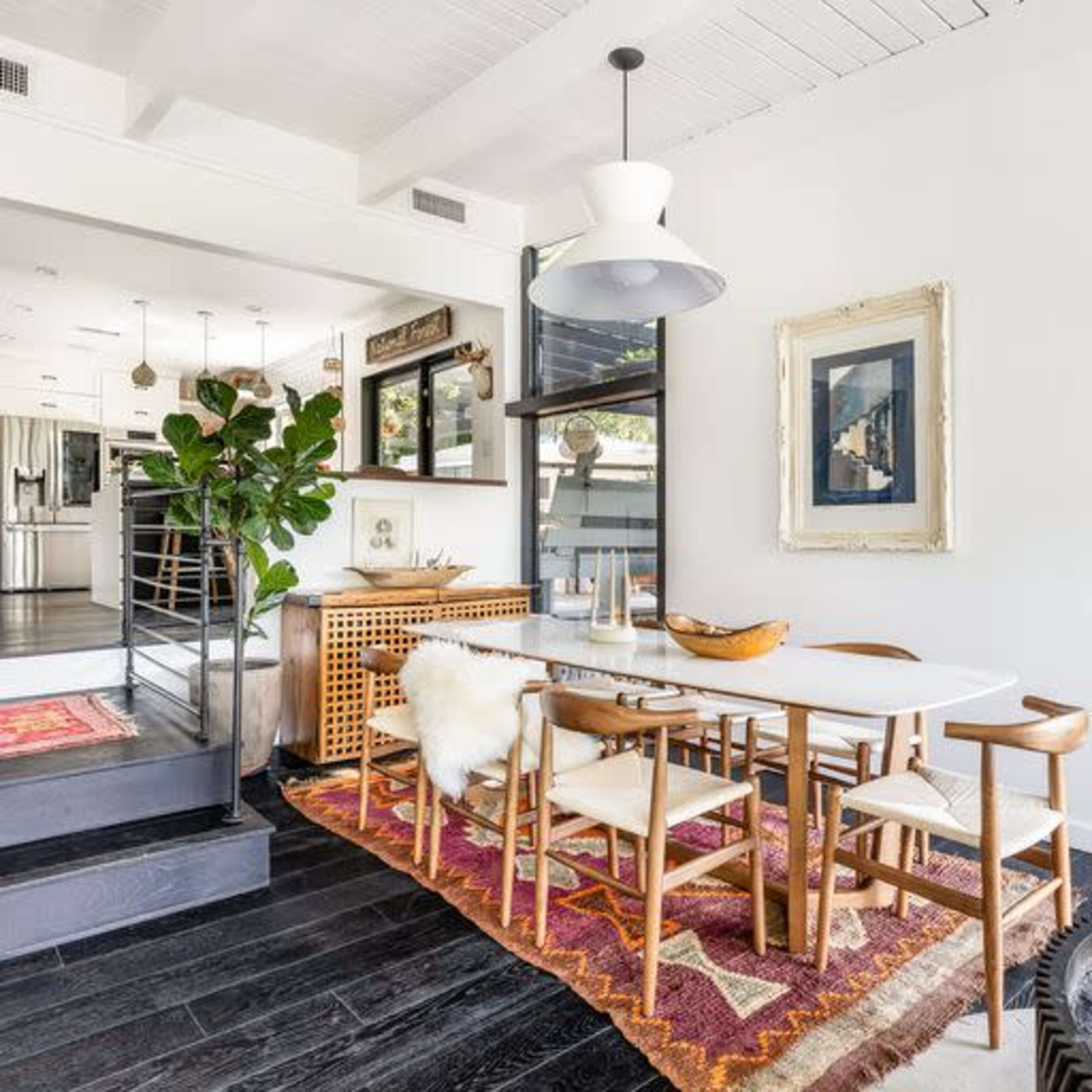 A modern dining area with a wooden table surrounded by chairs, a patterned rug on the floor, and a large window letting in natural light.