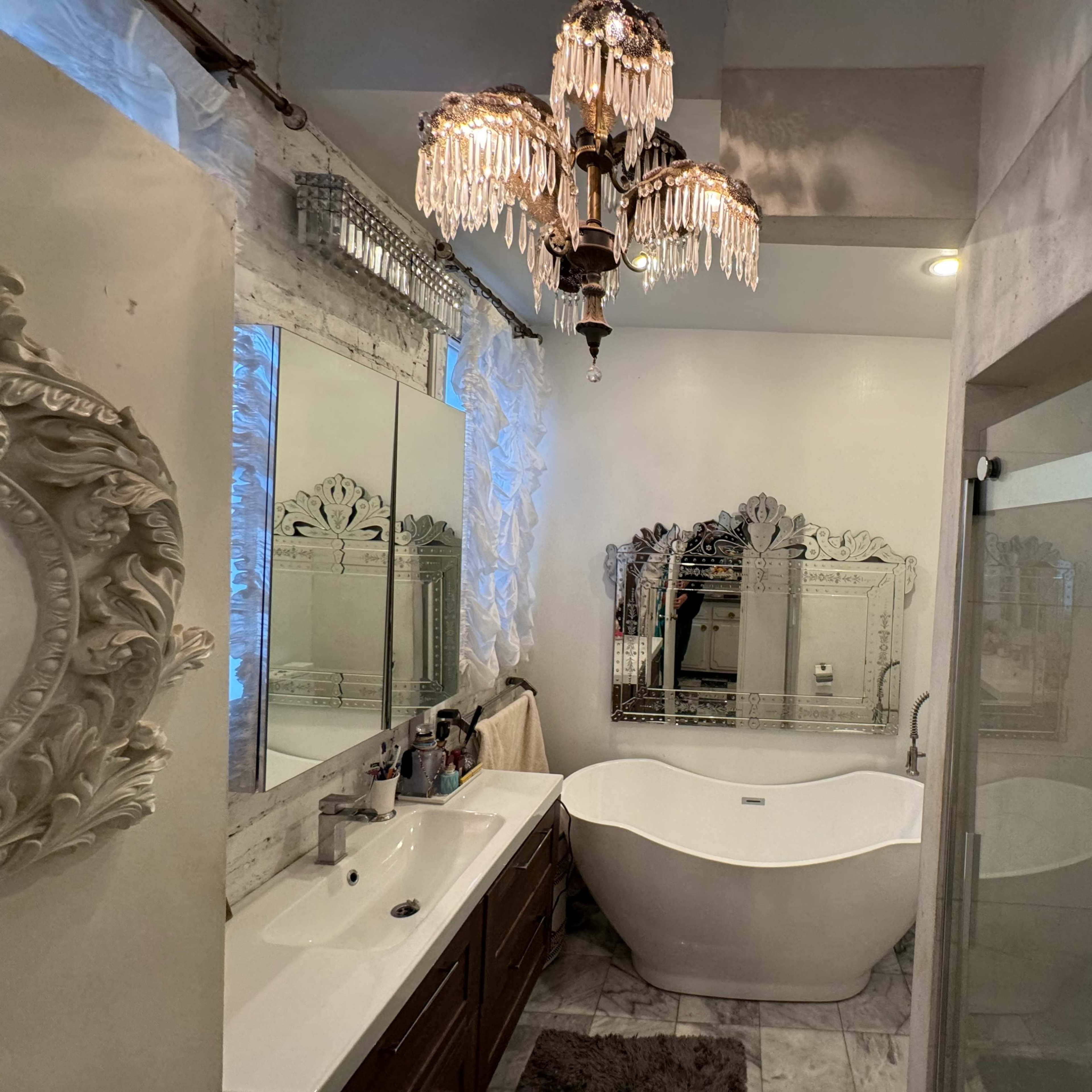 A modern bathroom featuring a freestanding bathtub, a large mirror with ornate detailing, and a chandelier hanging from the ceiling.