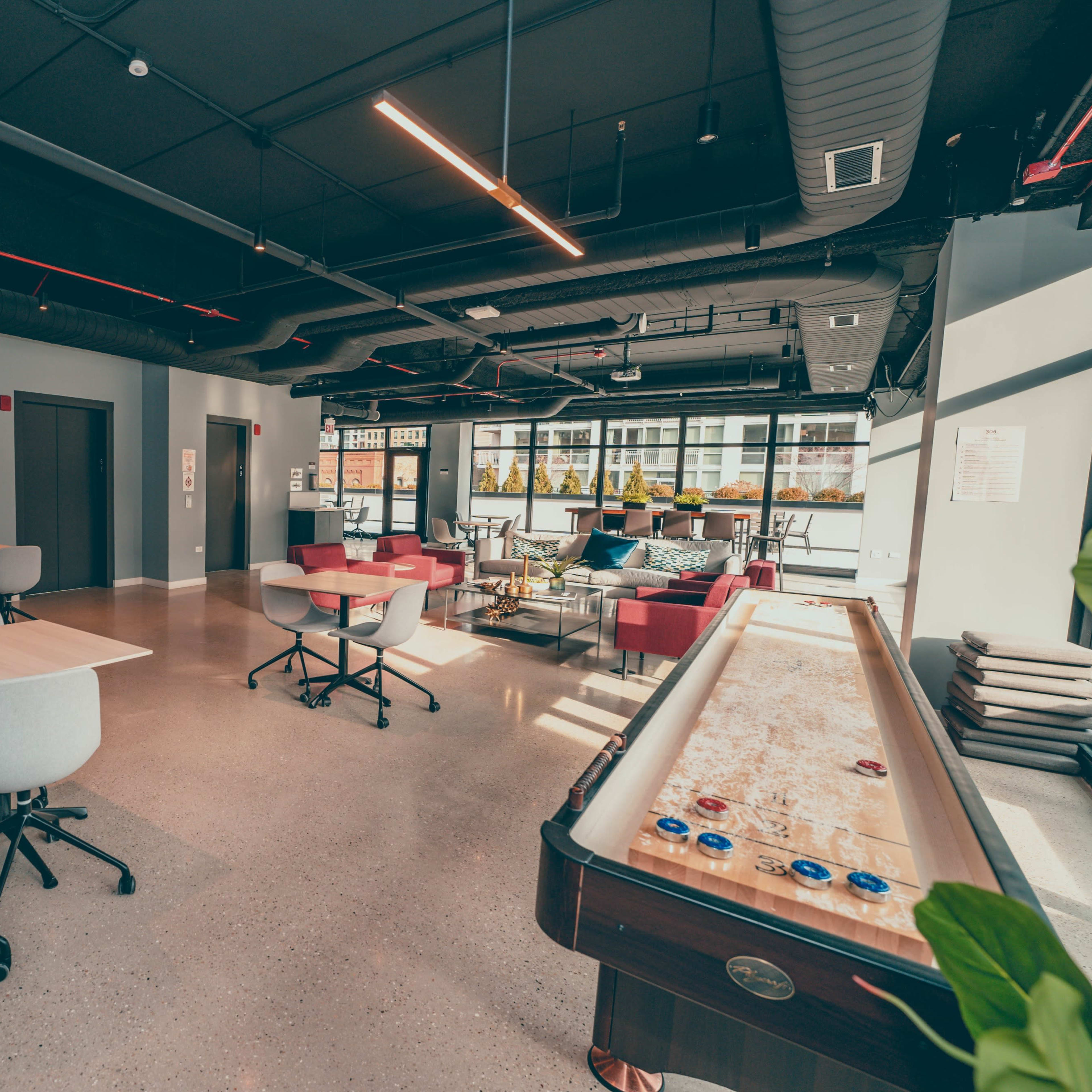 A modern communal workspace featuring tables, chairs, and a game area with a shuffleboard table, illuminated by natural light from large windows.