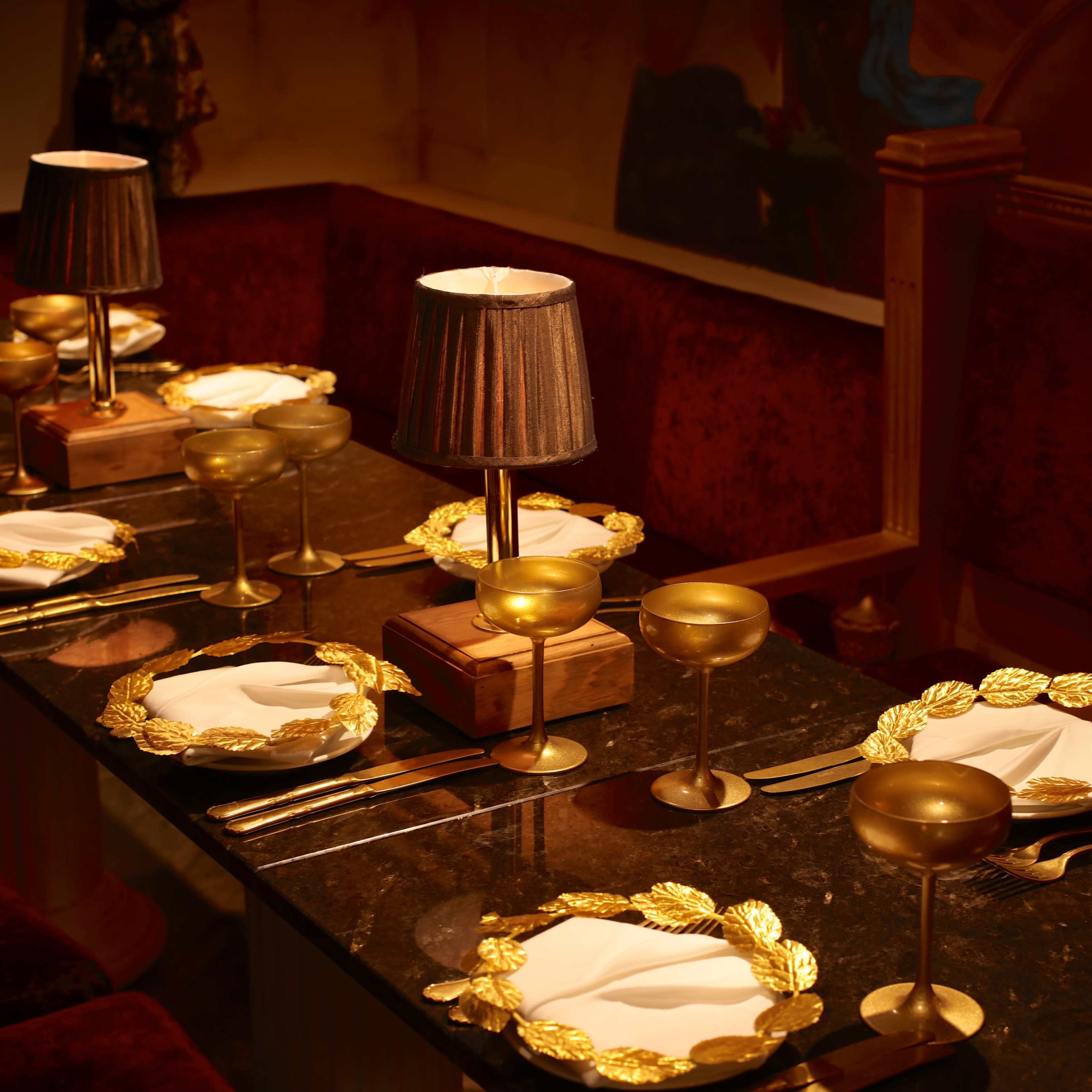 A long table is set with golden garnished plates, crystal glasses, and lamps, surrounded by velvet seating.